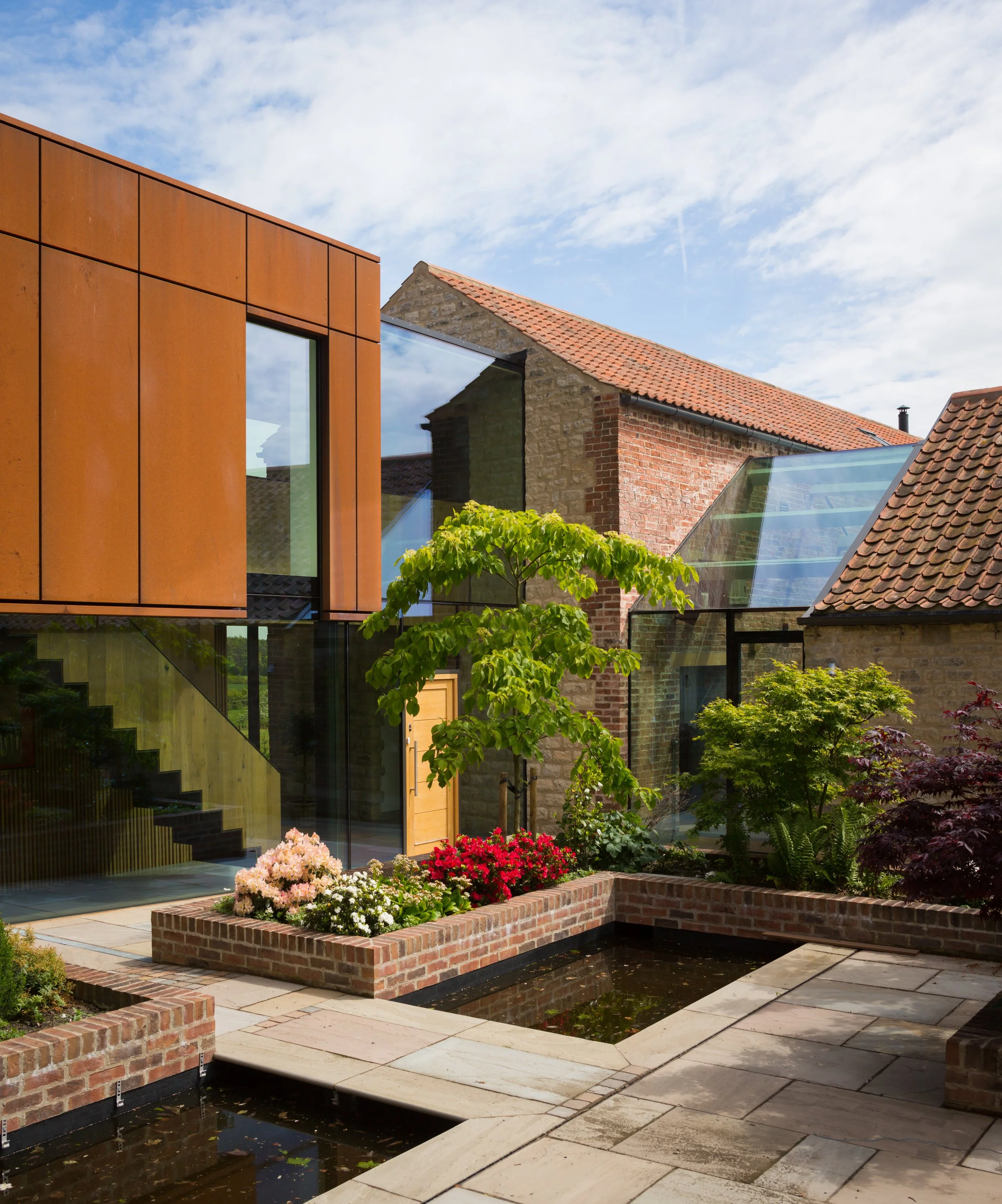 contemporary barn conversion, mixture of tall glass panelling and raw metal cladding with a small pond and neat raised flower bed by the front door