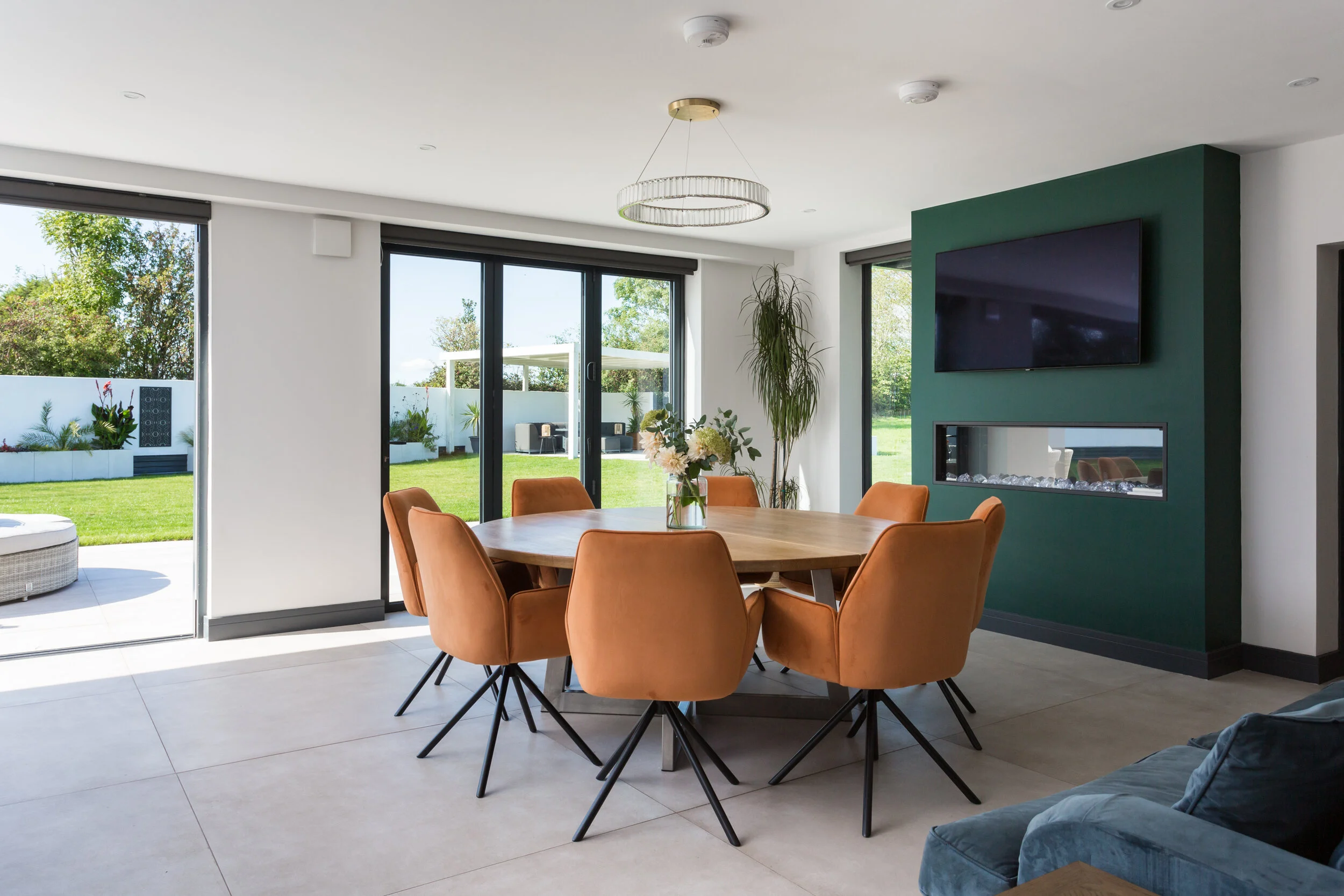 8 piece round dining table situated in a modern kitchen/diner next to large full height windows and a decorative fireplace painted forest green