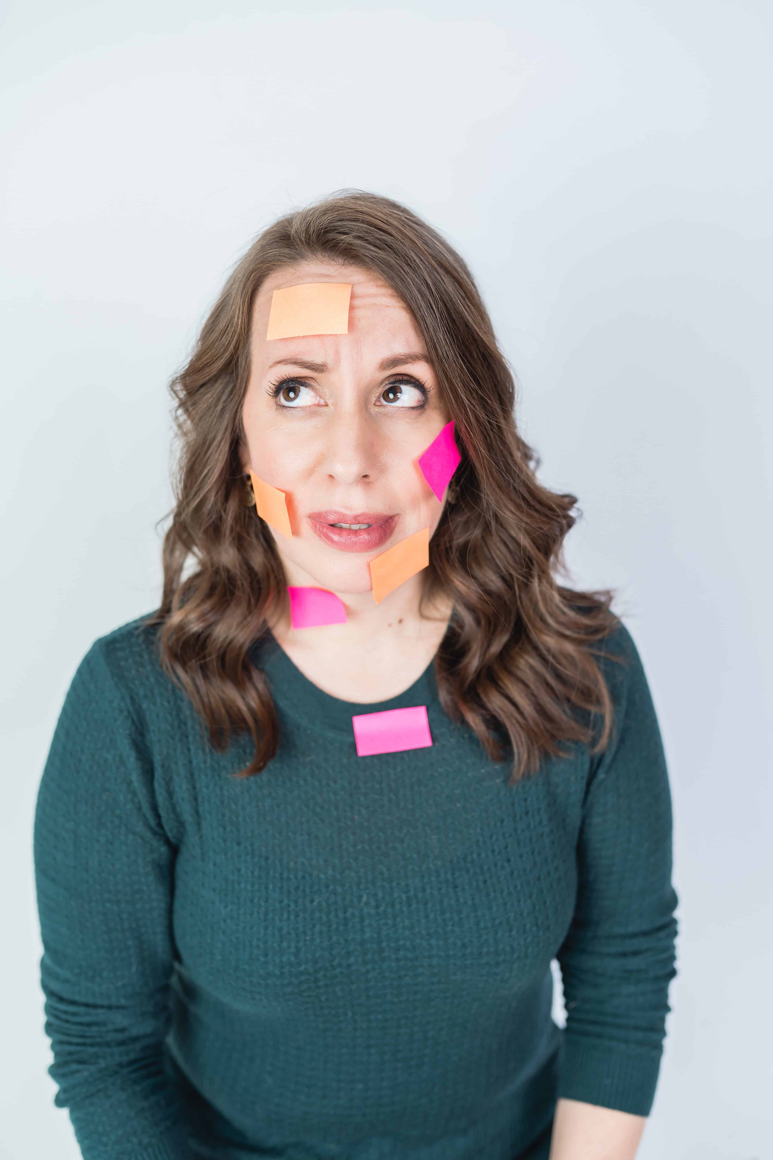 A woman with long brown hair wearing a green sweater, looking upwards with a curious expression, has multiple colorful sticky notes on her face and chest.