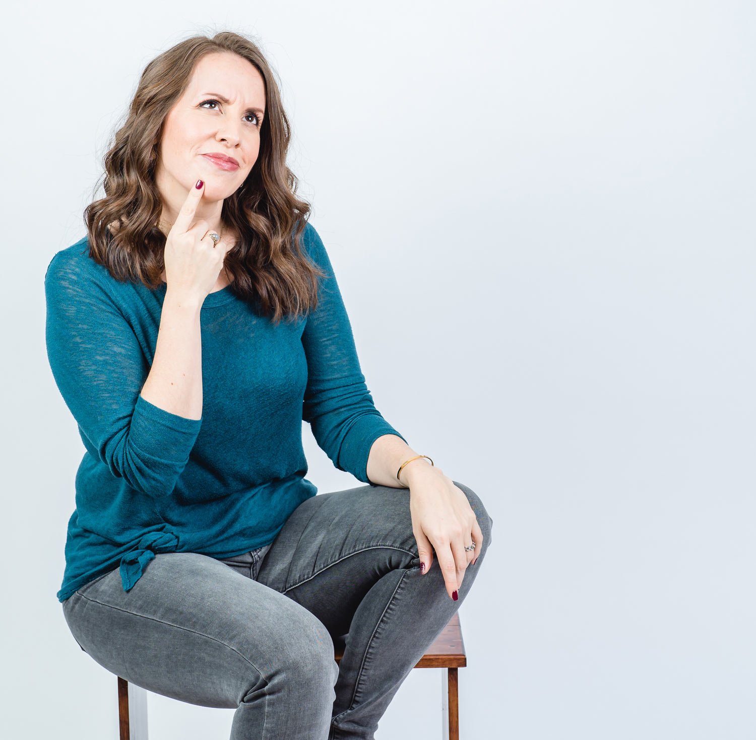 Woman in a blue sweater sitting on a stool, looking thoughtful with her hand on her chin.