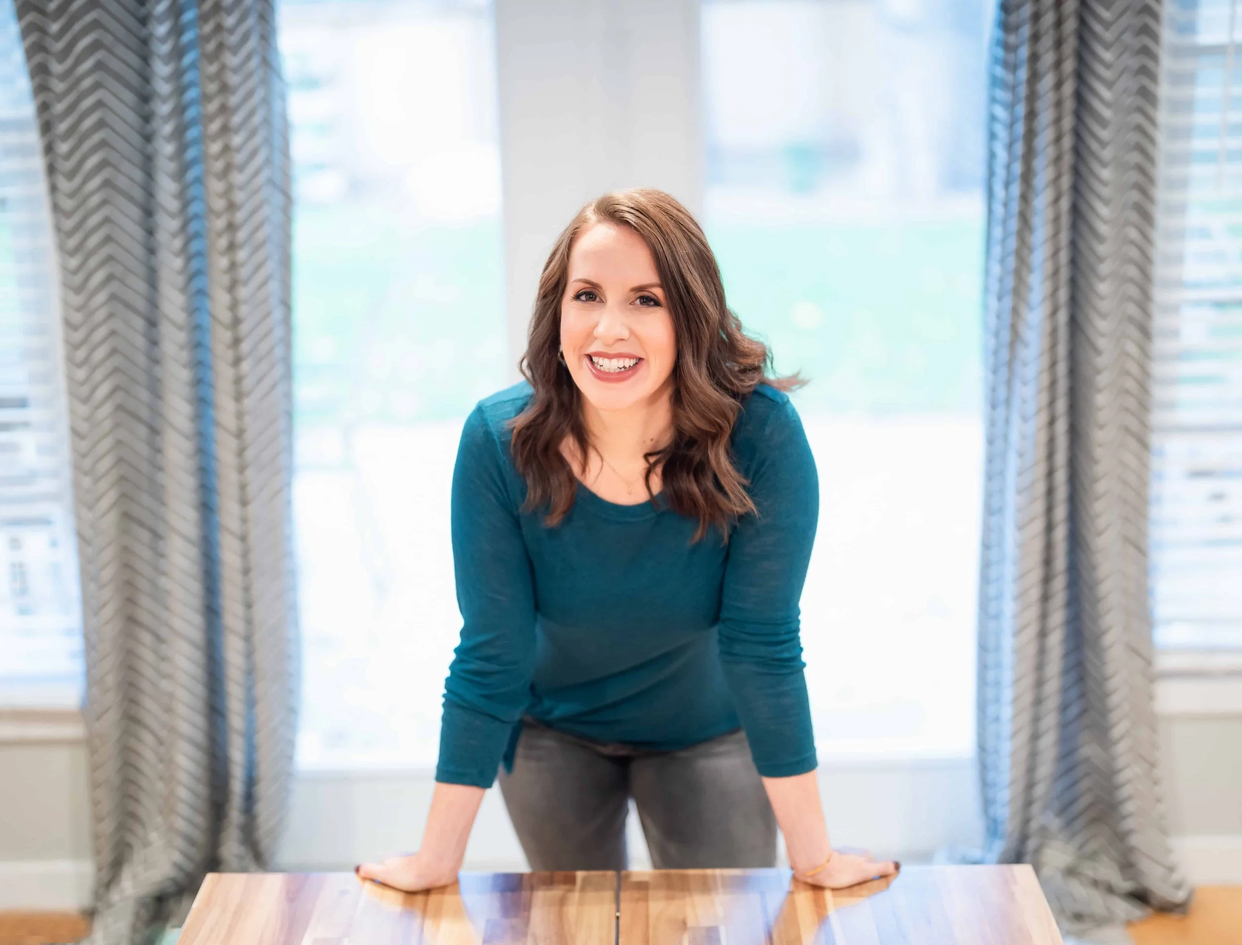 Smiling woman in a teal shirt leaning on a wooden table in a bright room with patterned curtains.