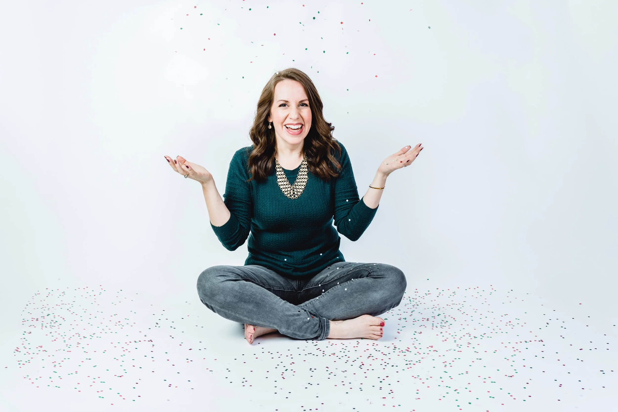 Woman sitting on floor with confetti, smiling, wearing green sweater and necklace, gray jeans, white background.