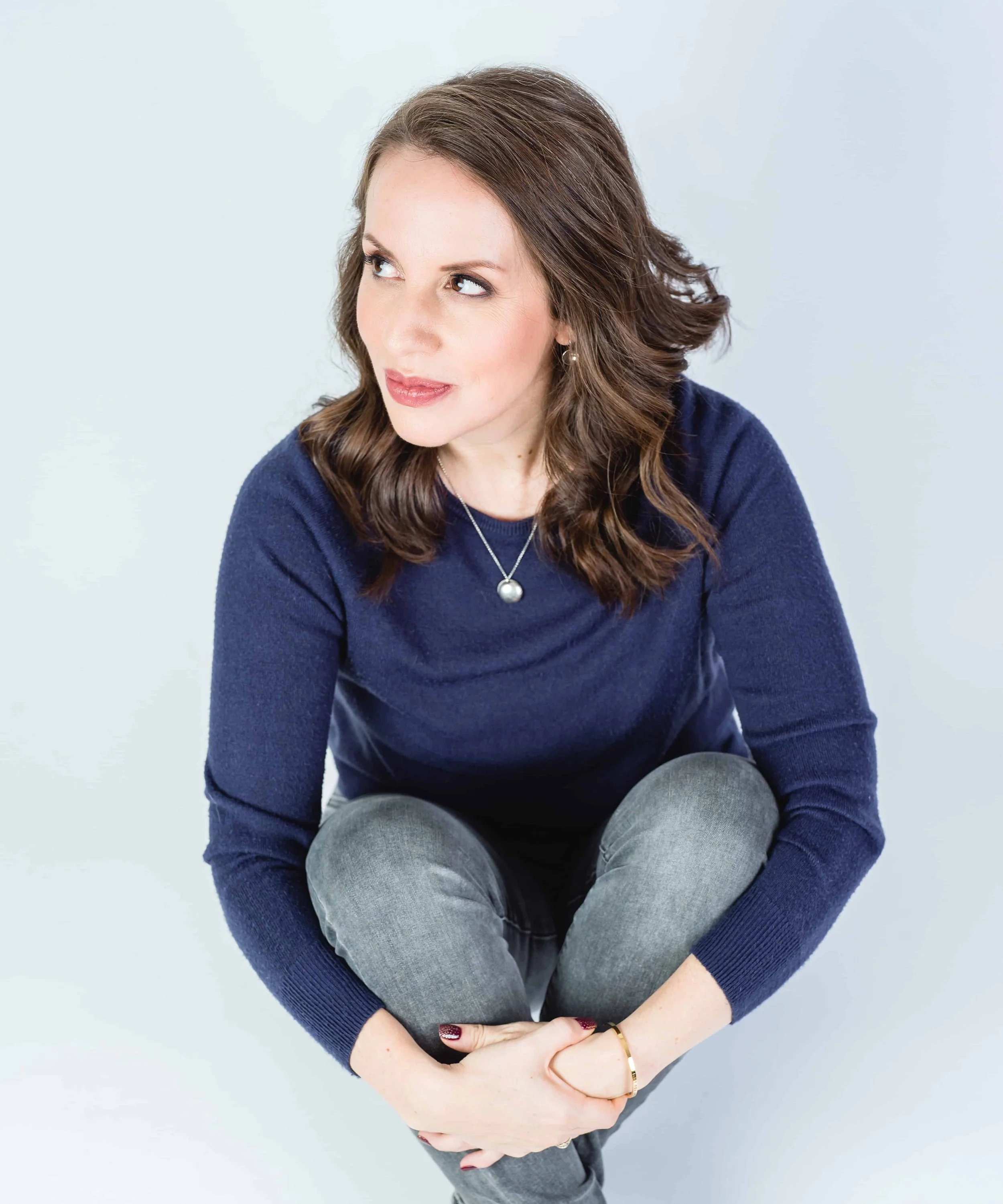 Woman wearing a navy sweater and gray jeans, sitting in a crouched position, looking off to the side, on a light background.