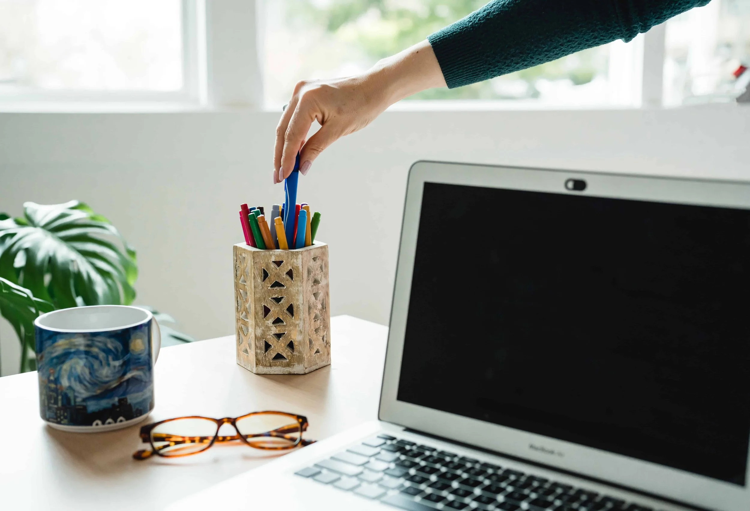 A hand reaching for a pen in a wooden holder, next to a laptop, coffee mug, and eyeglasses on a desk with a plant in the background.