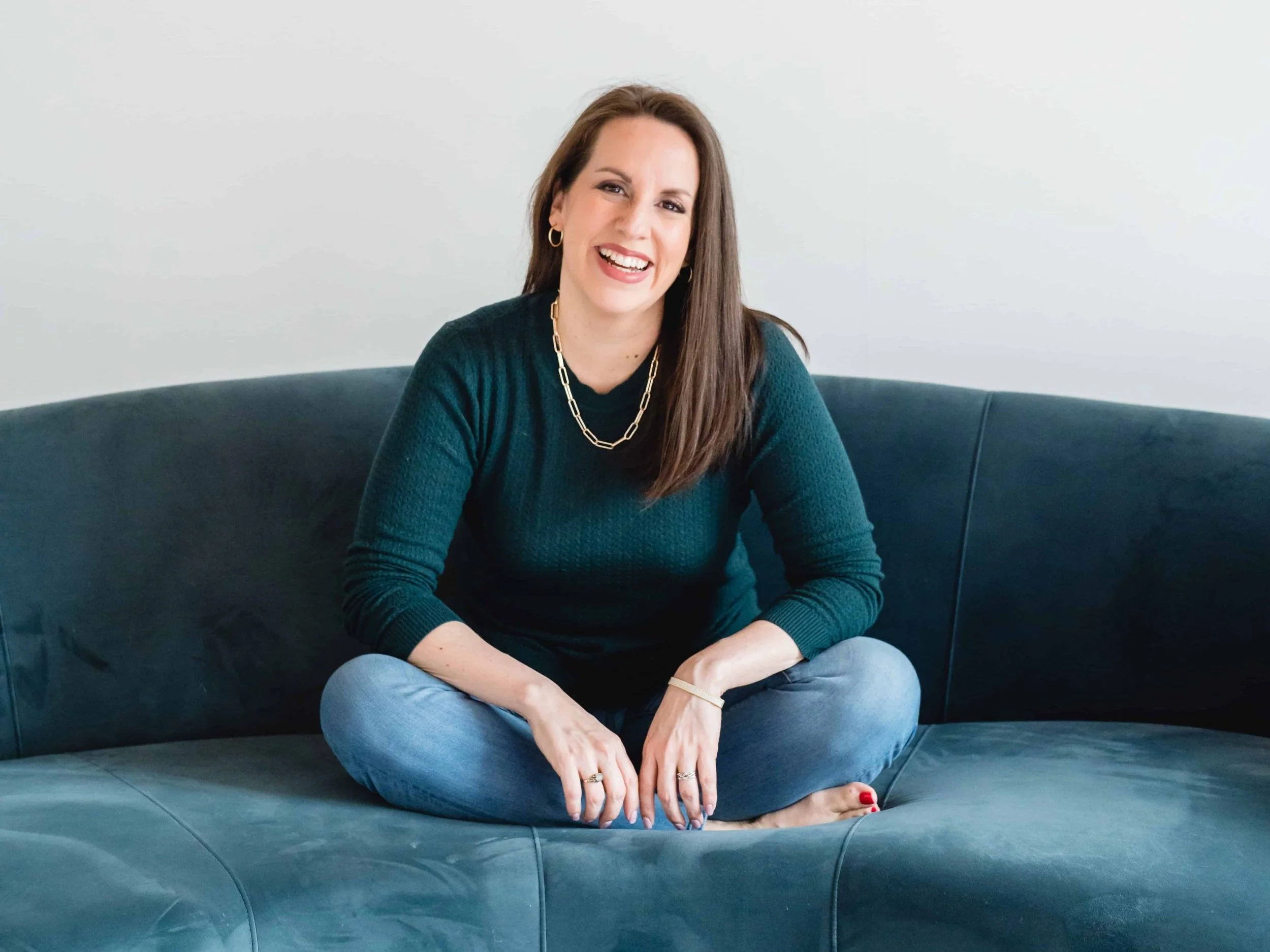A woman sitting cross-legged on a dark blue couch, wearing a dark green sweater and jeans, smiling at the camera against a plain background.