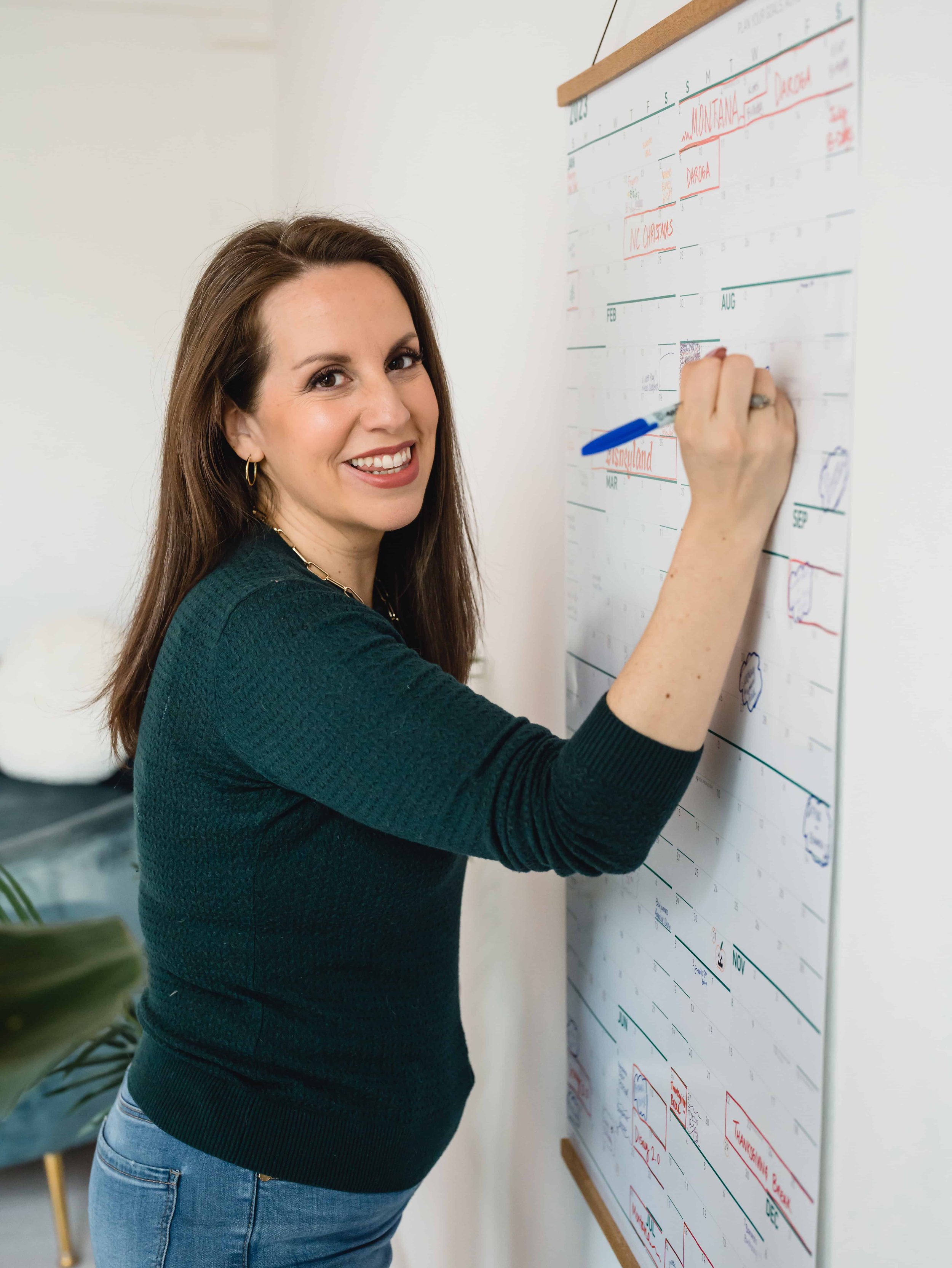 Woman with long brown hair smiling while writing on a wall calendar with a blue marker.