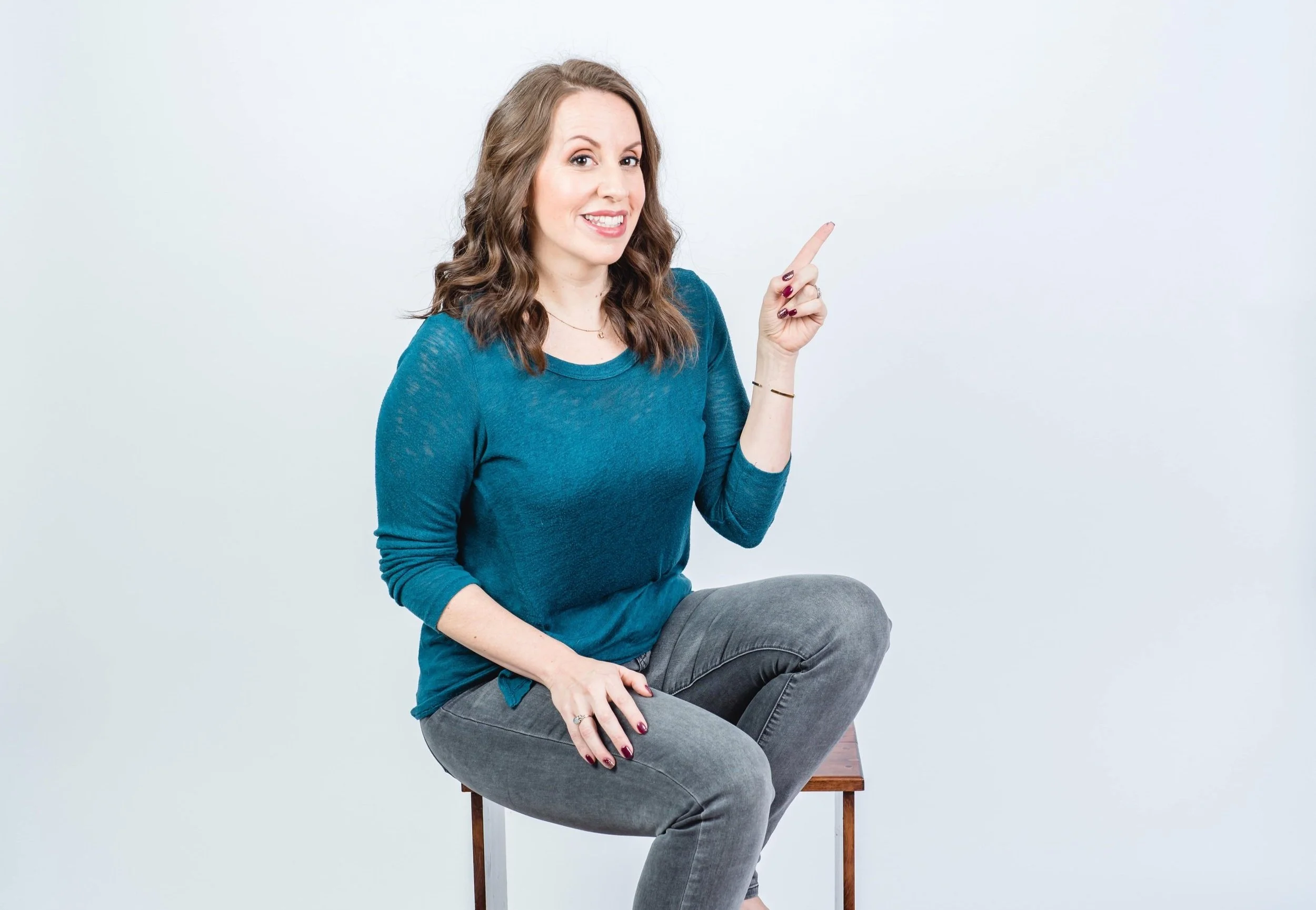Woman sitting on a stool, wearing a teal shirt and gray jeans, pointing upwards with her right hand, smiling.