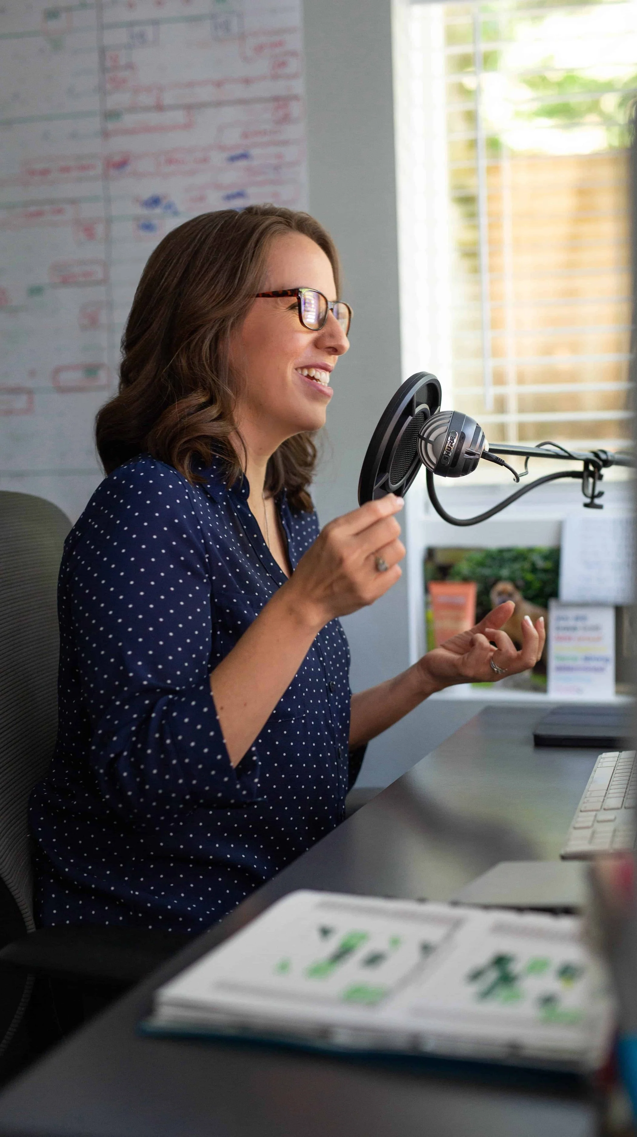 A woman with glasses and a blue polka dot shirt is speaking into a microphone on a desk in a well-lit room.