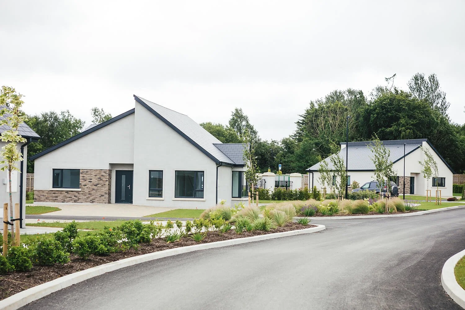 Modern white residential buildings with gray roofs, landscaped front yards, a curved asphalt driveway, small trees, and green shrubbery, in a suburban neighborhood.