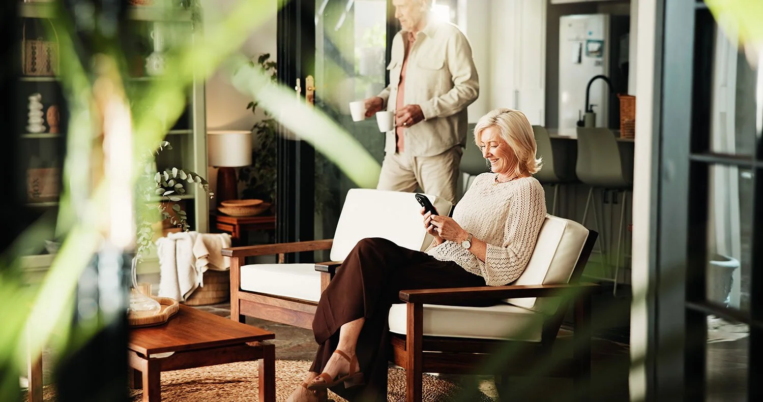A woman sitting on a cream-colored couch in a sunlit room, looking at her phone and smiling, while a man in the background holds coffee cups and stands near a door.