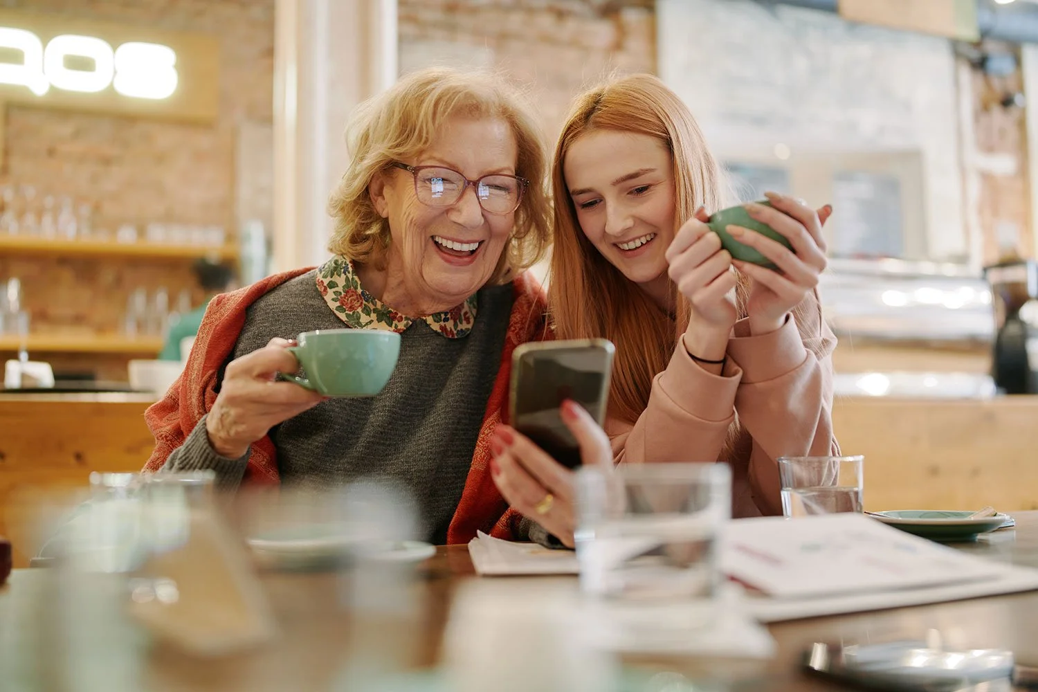 An grandmother and teenage grandaughter sharing a joyful moment in a coffee shop, looking at a smartphone together, with cups and glasses on the table.