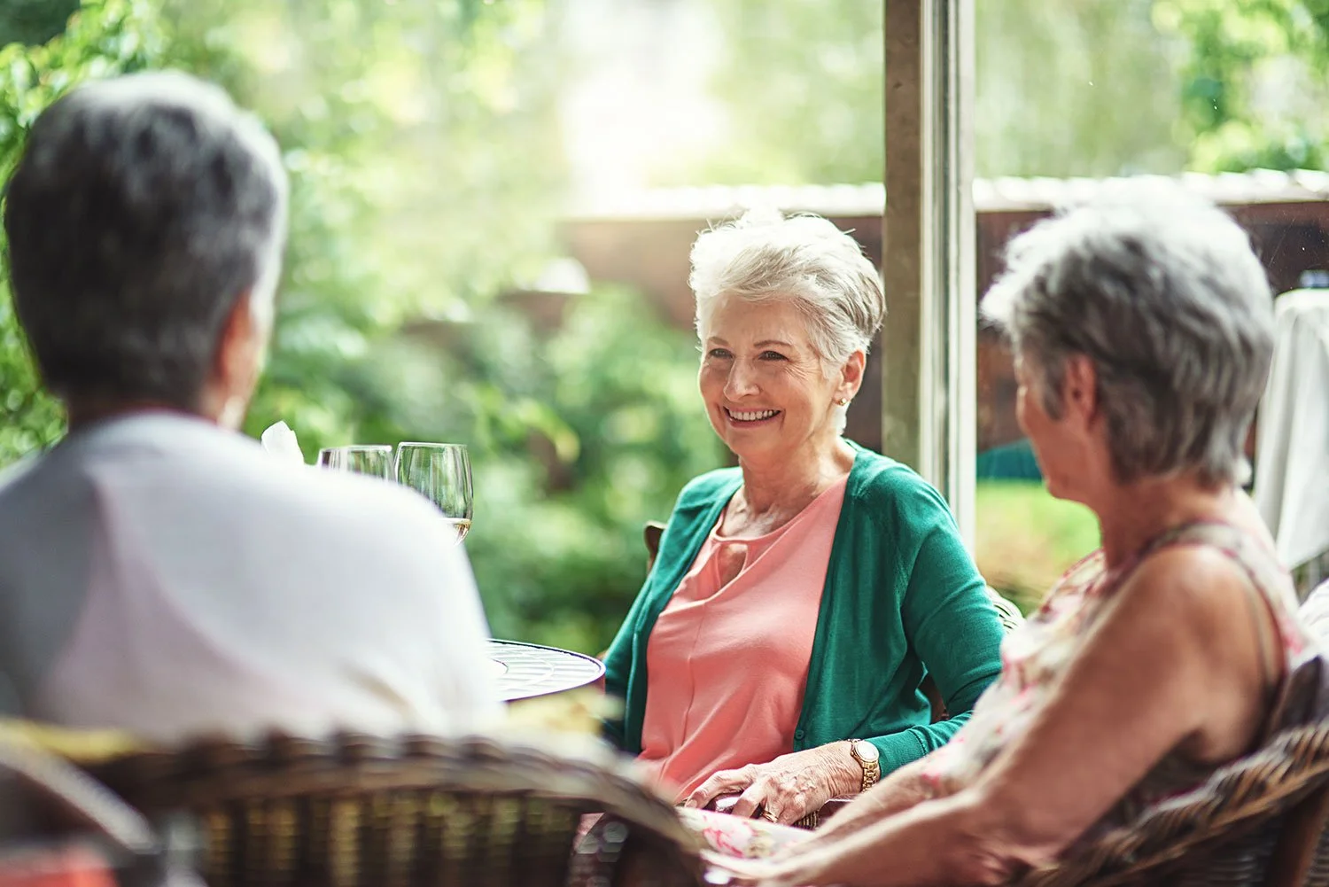 Three people sitting around a table, having a conversation outdoors with a green garden in the background. One woman in the center is smiling.