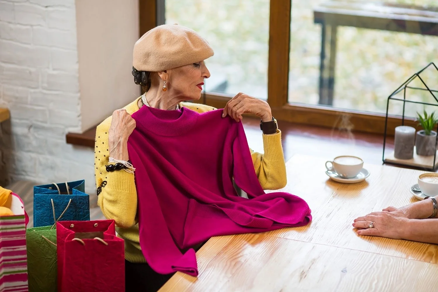 An older woman with earrings and bracelets, wearing a tan beret and colorful sweater, sits at a wooden table holding a pink sweater. There are gift bags on the table and tea cups nearby, with a window in the background showing autumn scenery.