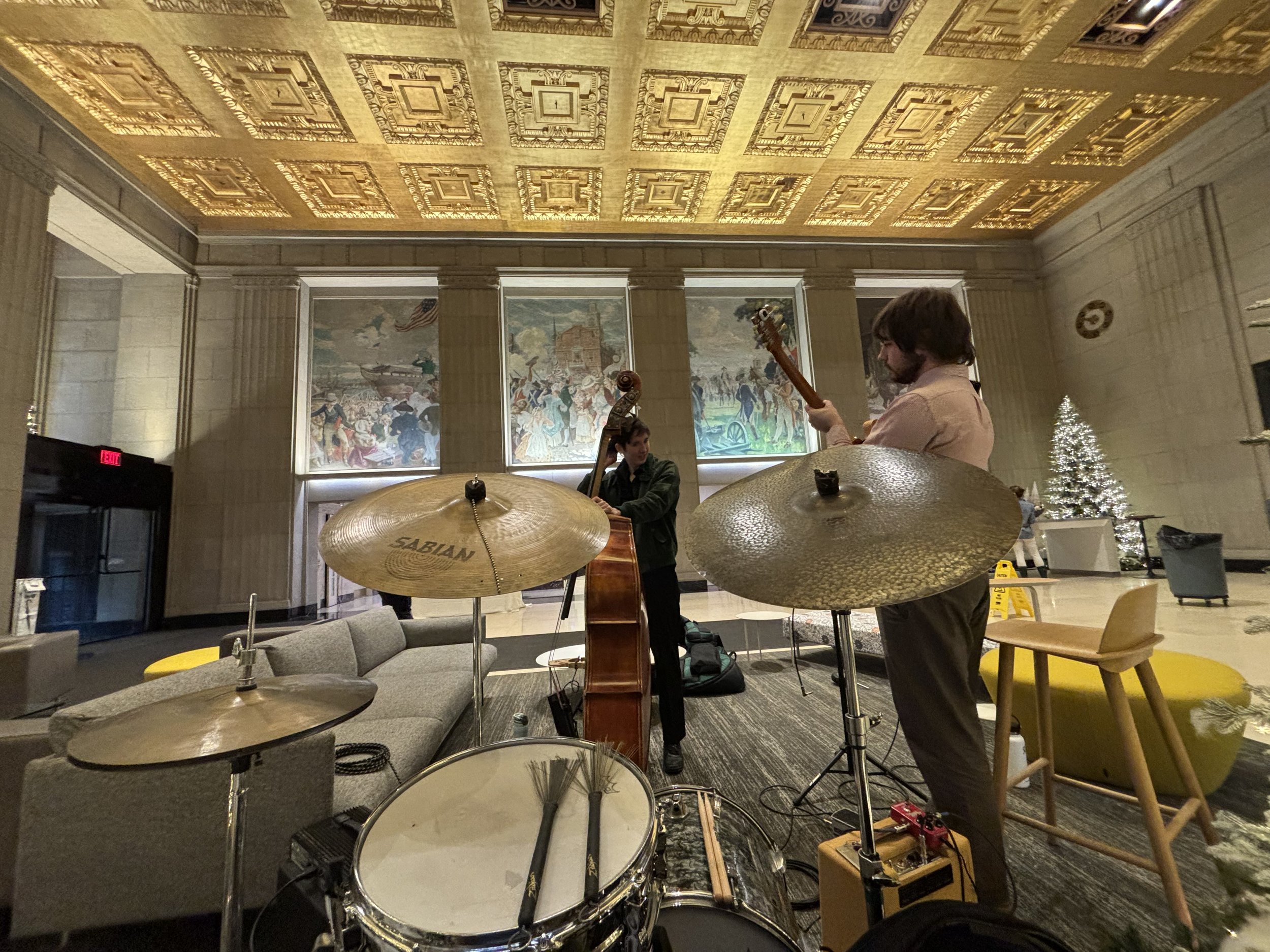 Guitarist and upright bassist performing beside a drum kit and illuminated Christmas trees at 501 Boylston