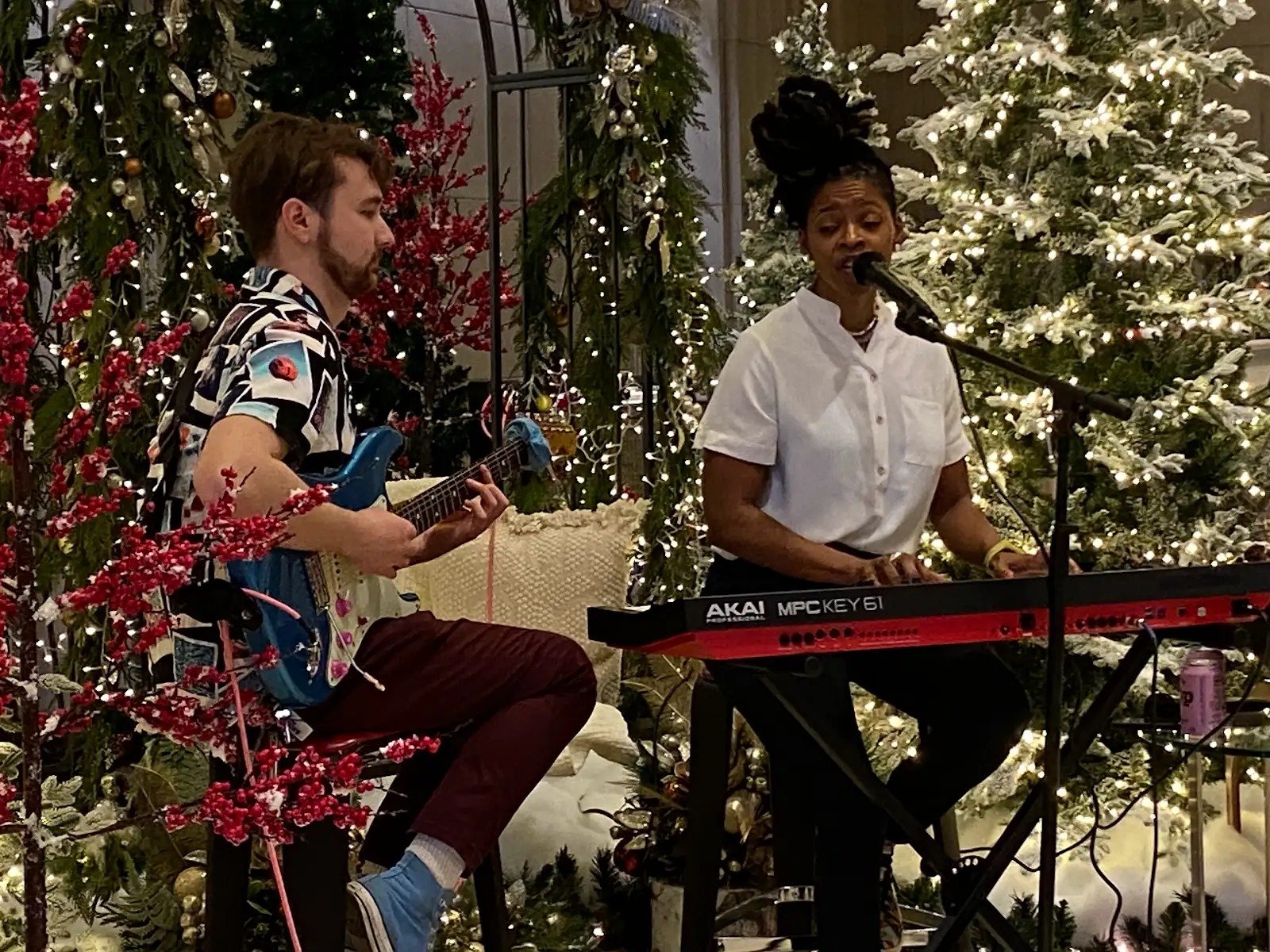 Vocalist at a keyboard and guitarist performing beside decorated Christmas trees at a holiday event