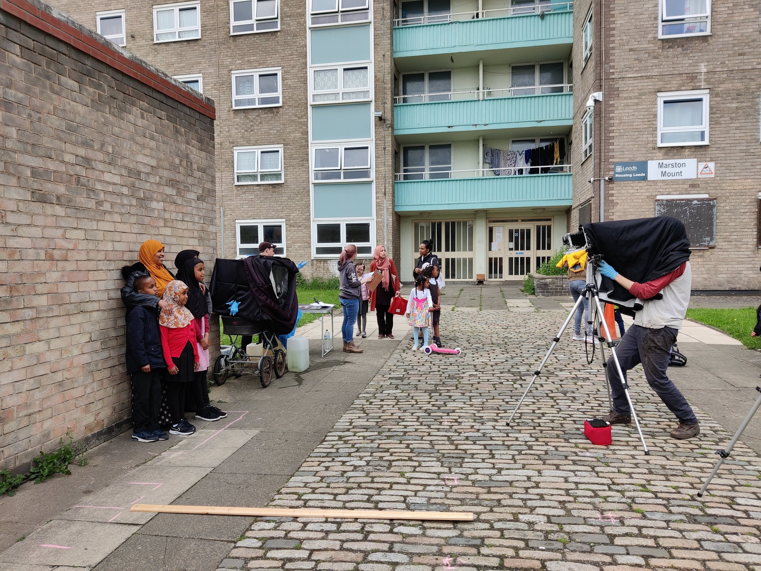 A group of children and adults standing outside on a cobblestone street near a brick building, with a camera and crew filming them.