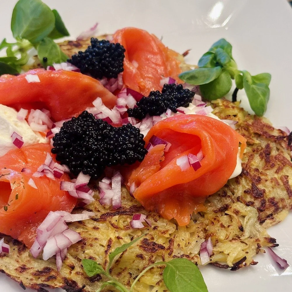 A golden, crispy potato rösti topped with ribbons of pink gravlax, dollops of crème fraîche, black caviar, diced red onions, and fresh green microgreens.