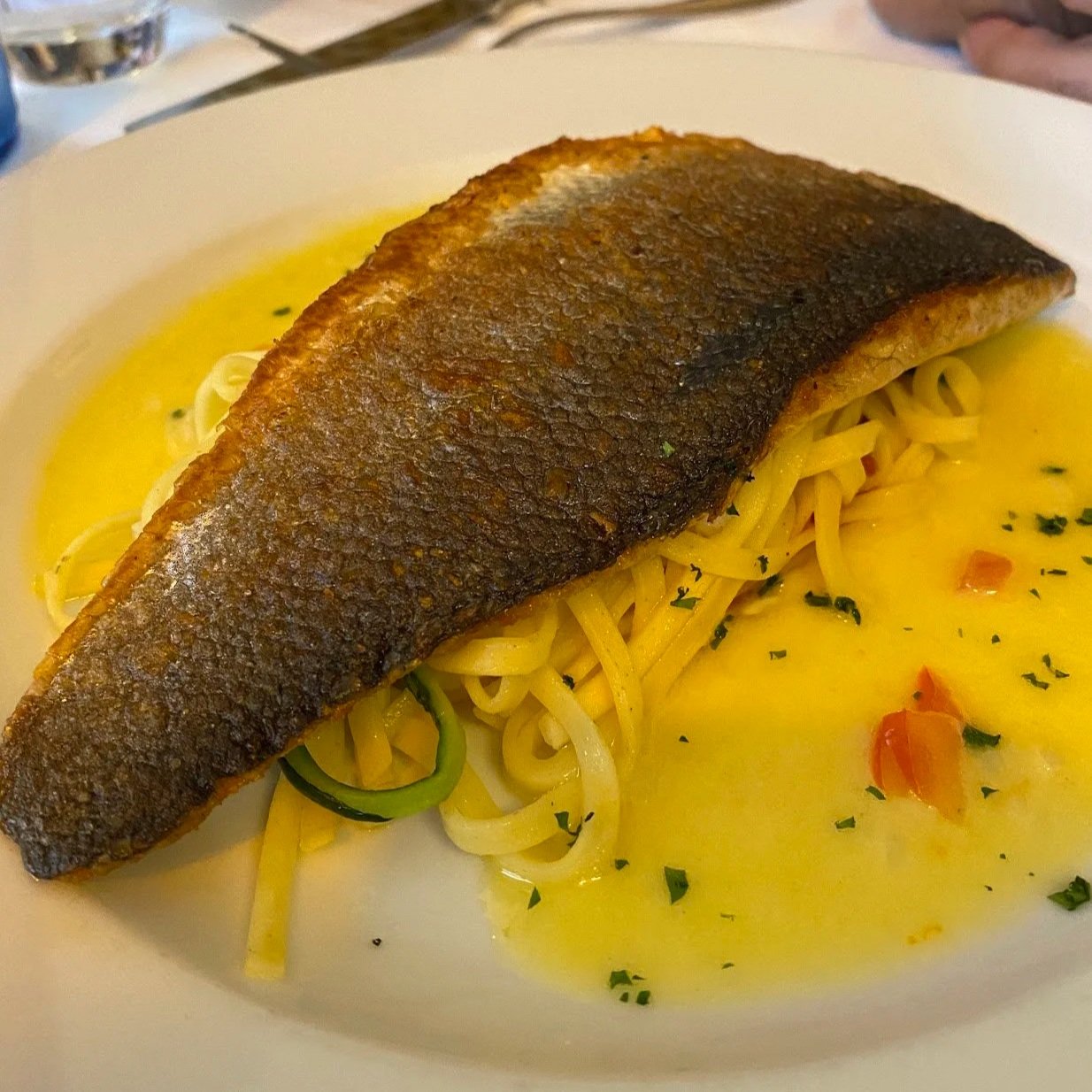 A crispy skin-on sea bass fillet served over a nest of linguine pasta, surrounded by a bright yellow beurre blanc sauce with diced tomatoes and herbs.