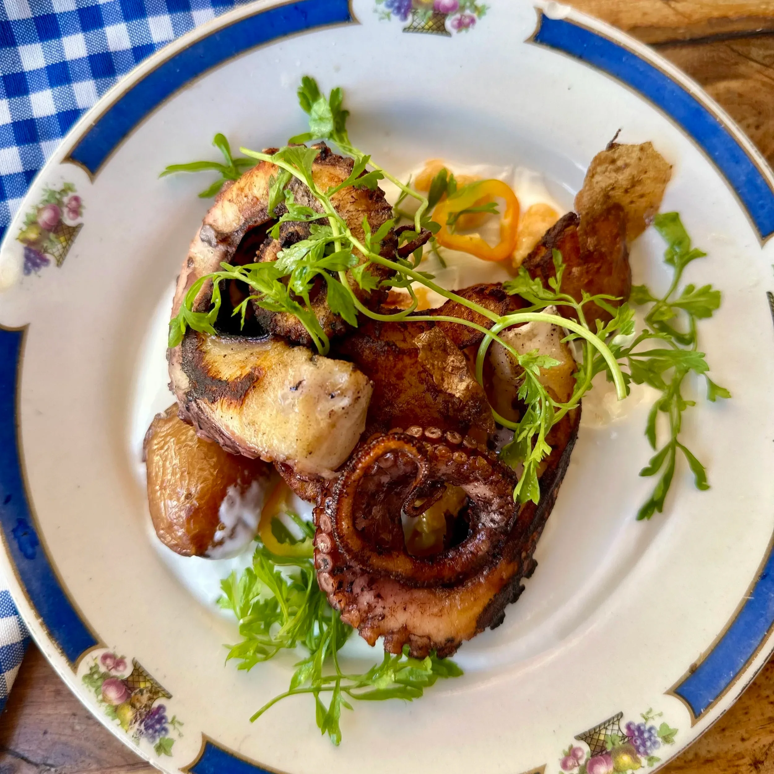 charred octopus tentacles and golden smashed potatoes. The components sit atop a creamy white yogurt base, garnished with bright green chervil sprigs and yellow chili rings, served on a vintage blue-rimmed floral plate.