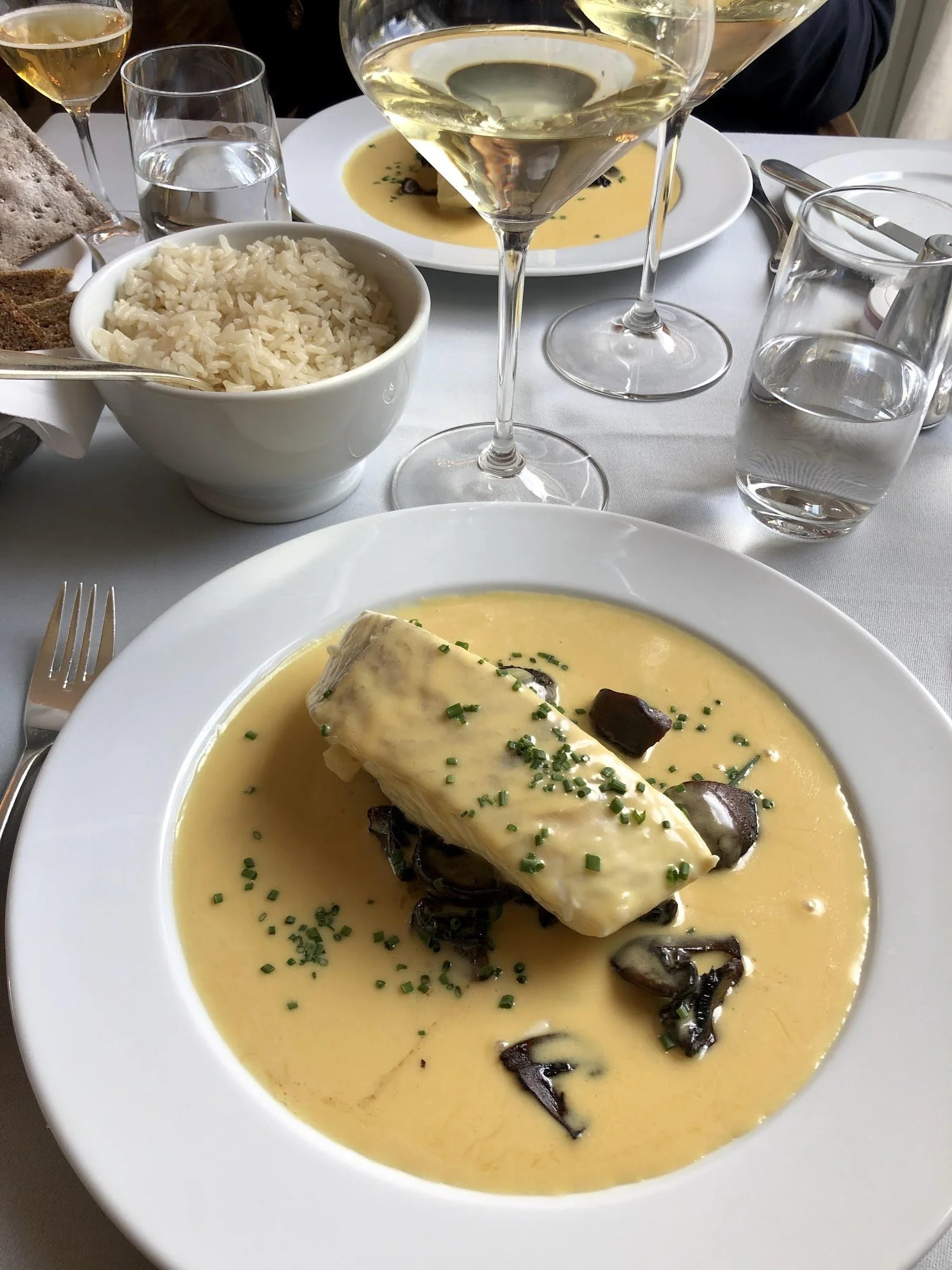 A thick, white poached cod loin draped in a pale yellow champagne sauce, garnished with chives and mushrooms, served with a side of white rice.