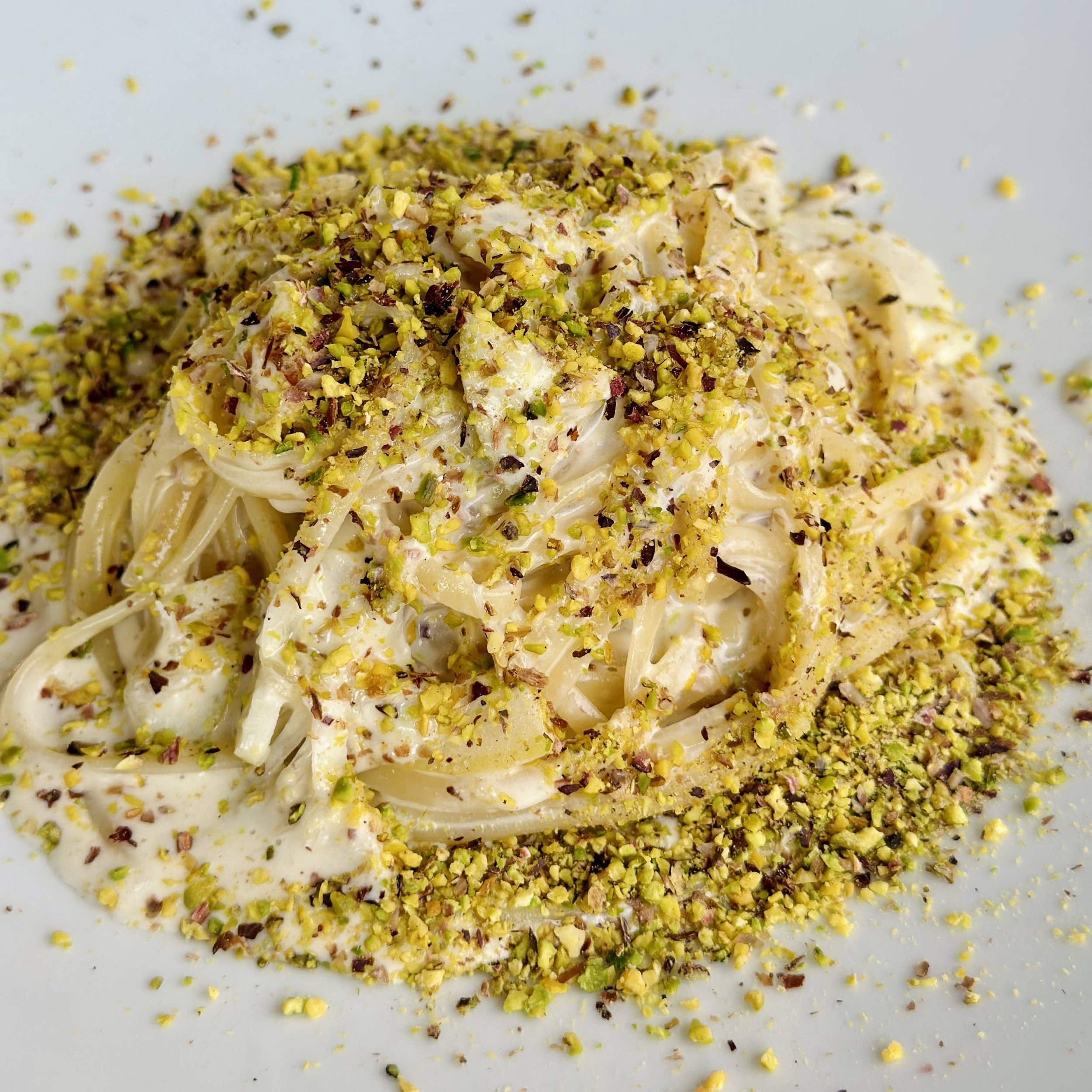 A high mound of creamy, white-sauced linguine pasta on a white plate, heavily showered with finely chopped green and yellow pistachios.