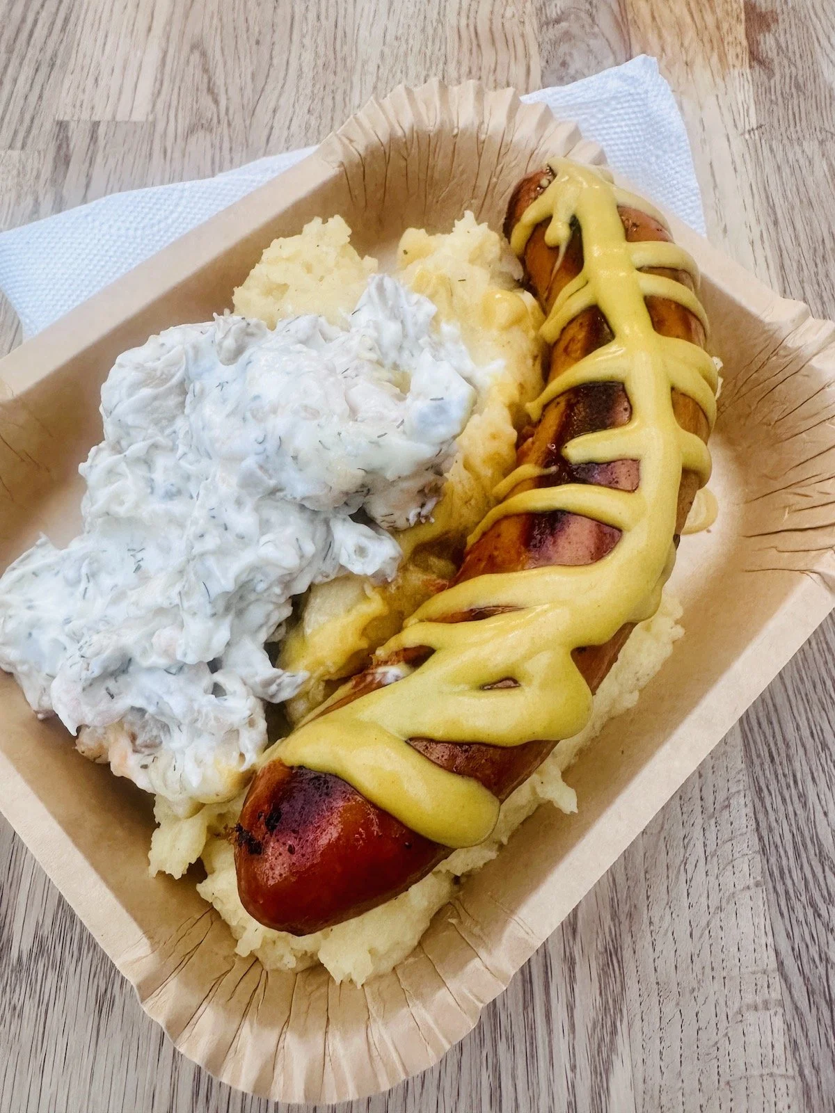 Grilled chorizo on a bed of mashed potatoes in a paper tray, topped with a zig-zag of yellow mustard next to creamy shrimp salad.
