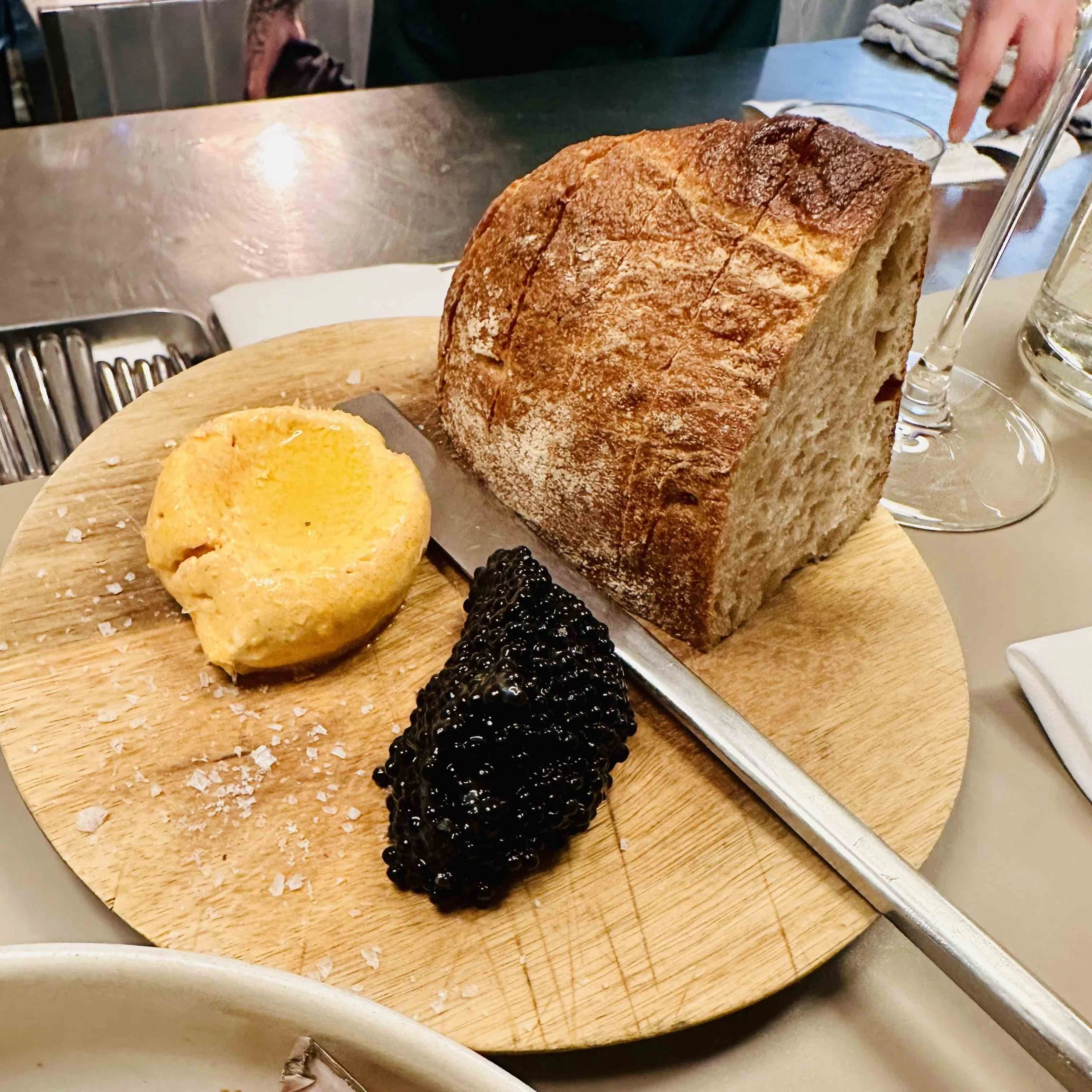 A thick, crusty slice of golden-brown sourdough bread served on a wooden board alongside a mound of creamy yellow horseradish butter and a portion of glistening black seaweed caviar.