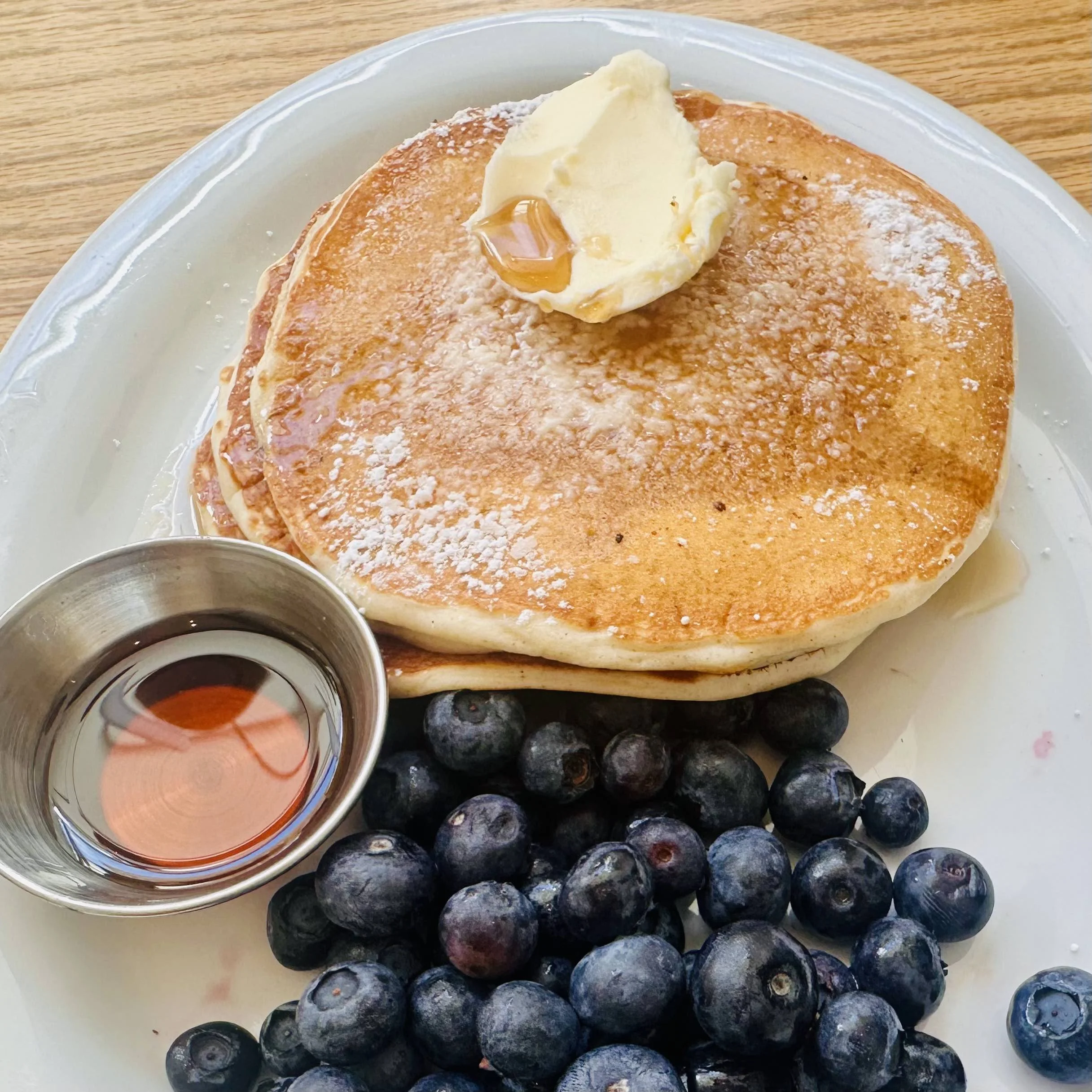 A stack of golden pancakes topped with butter and powdered sugar, served on a plate with a side of maple syrup and fresh blueberries.
