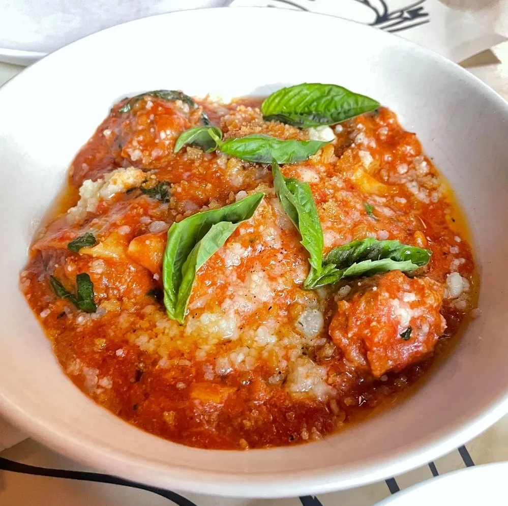 A white bowl filled with tender meatballs simmered in a bright red tomato sauce, topped with grated cheese and vibrant fresh basil leaves.