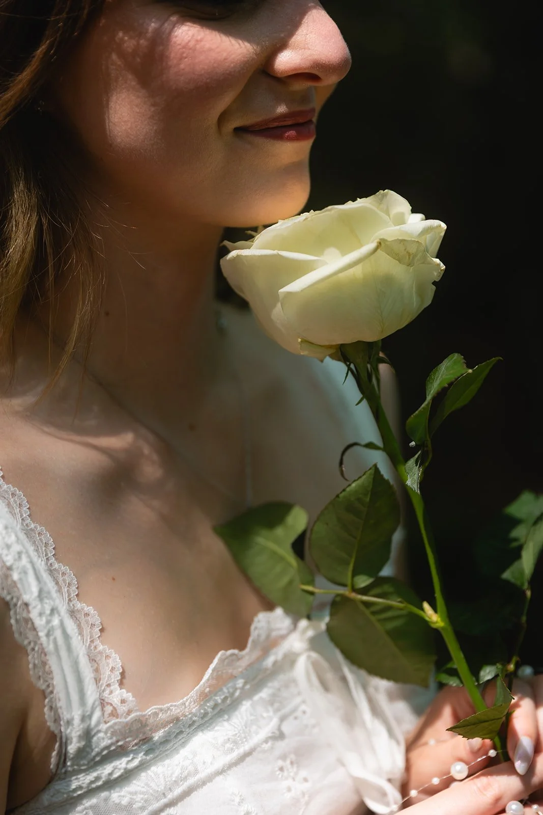 A woman holding a white rose near her face, dressed in a white lace top, with a dark background.