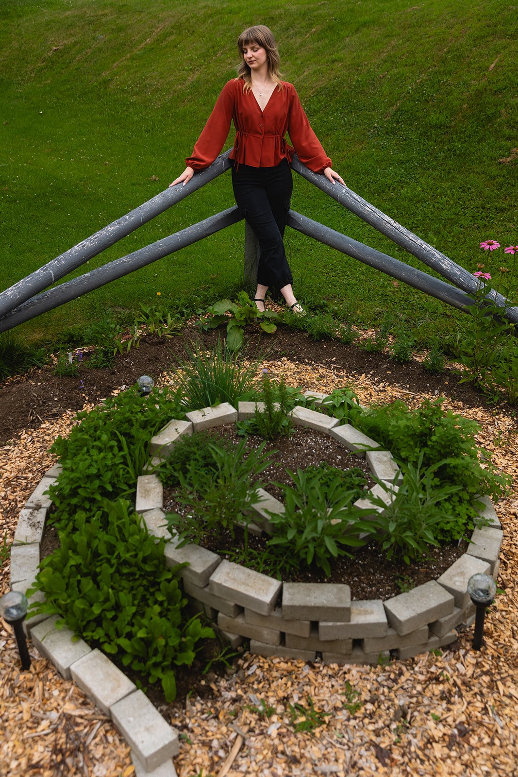 A woman with shoulder-length brown hair wearing a red blouse and black pants, leaning against a wooden railing in a garden with green grass and a flower bed with small green plants and flowers.