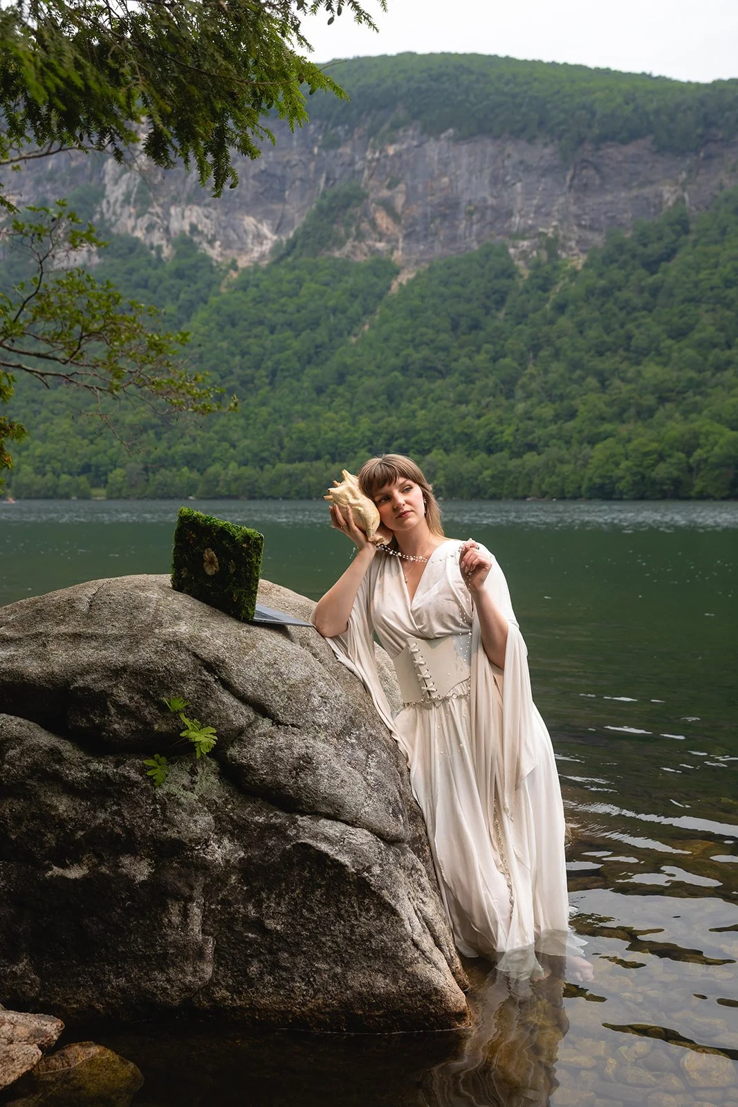 A woman in a vintage-style white dress sitting on a large rock by a lake, holding a conch shell to her ear, with a laptop and a moss-covered book nearby, surrounded by lush green trees and mountains.