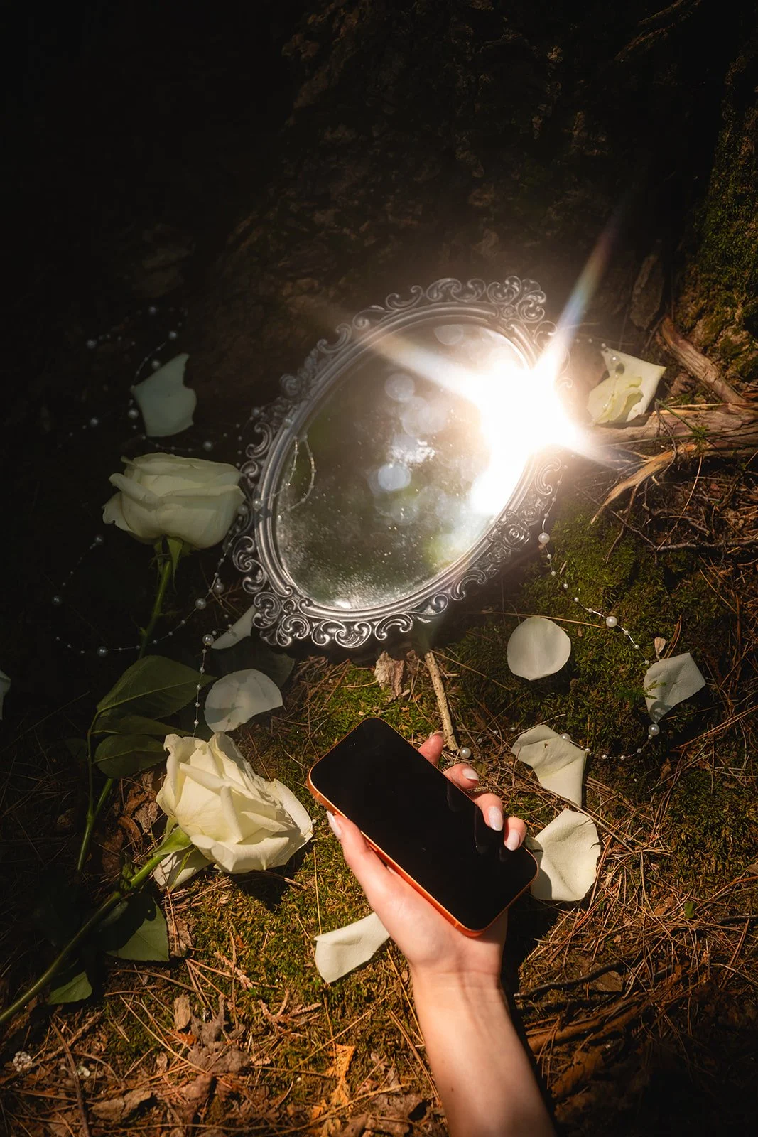 An ornate mirror on the ground surrounded by white roses, flower petals, and a string of pearls, with a hand holding a smartphone nearby, in a dark outdoor setting.
