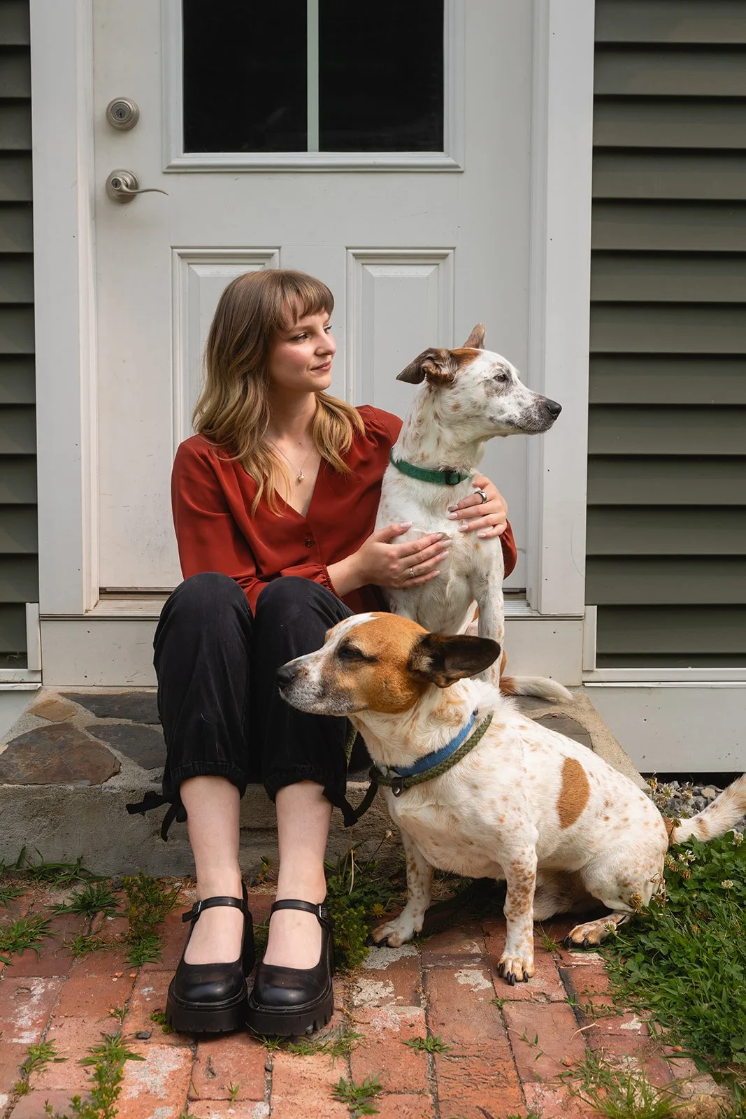 A woman sitting on a brick porch with two dogs, one sitting on her lap and the other sitting next to her. She is wearing a red blouse and black pants. The background features a white door with a window. The setting appears to be outside a house.