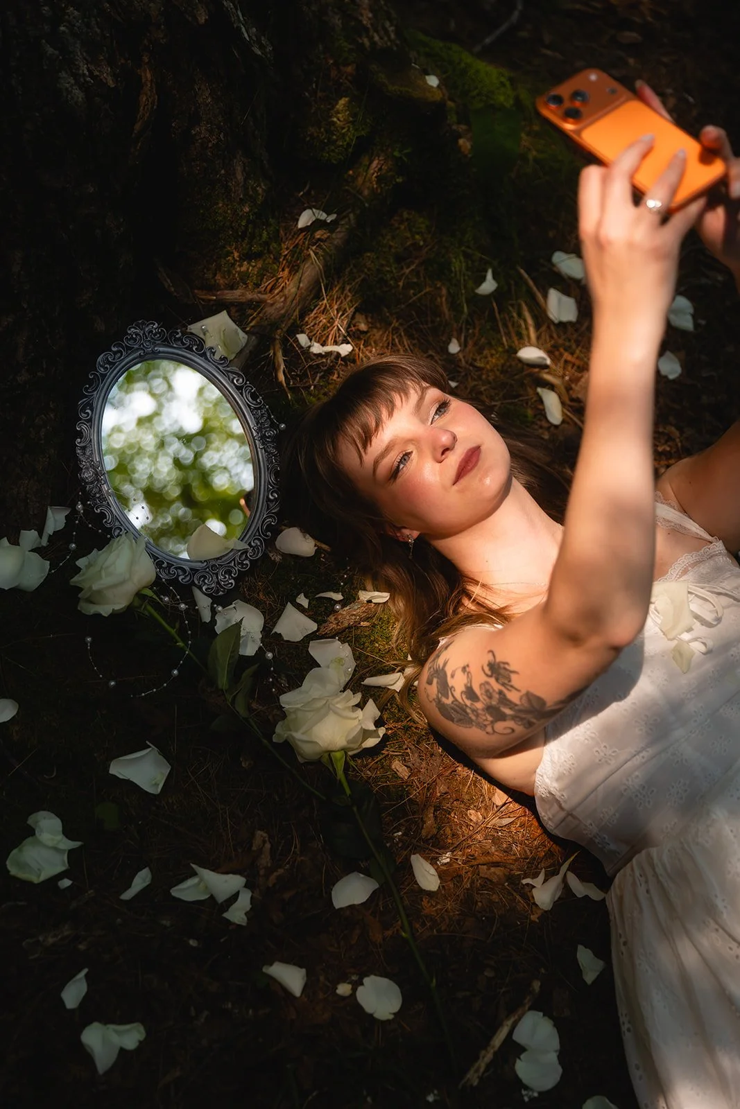 A woman lying on the forest ground surrounded by white flower petals, taking a selfie with an orange phone, with a small mirror reflecting trees and sunlight.