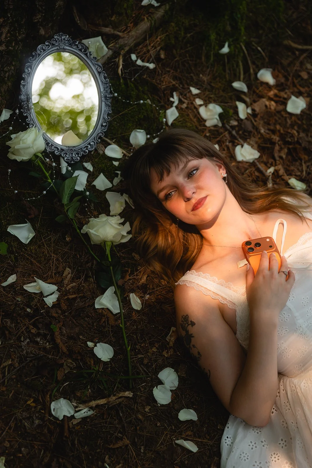 A young woman with brown hair lying on the ground among white rose petals and leaves, holding a phone, with a decorative mirror nearby reflecting a blurry outdoor scene.