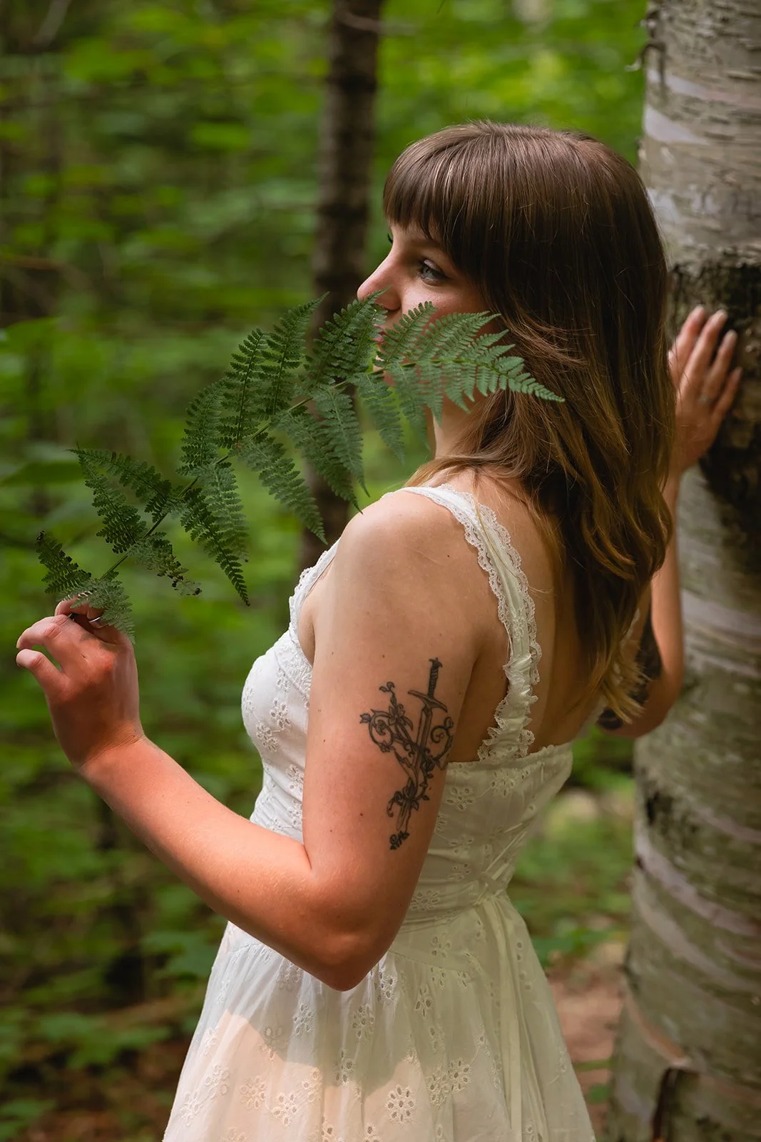 A woman with shoulder-length brown hair and a tattoo on her upper arm stands in a forest, holding a fern leaf in front of her face, with her right hand touching a tree trunk.