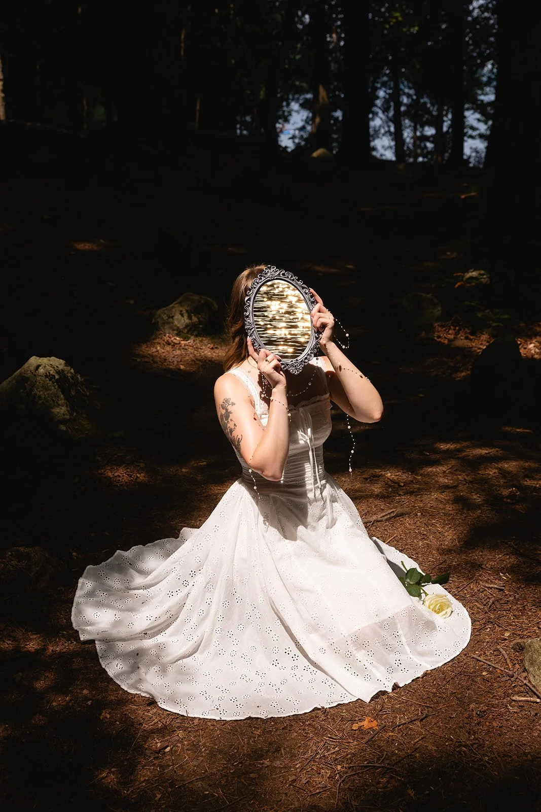 A woman in a white wedding dress kneels on the forest floor holding an ornate mirror in front of her face, reflecting water and sky. A white rose lies on the ground beside her.