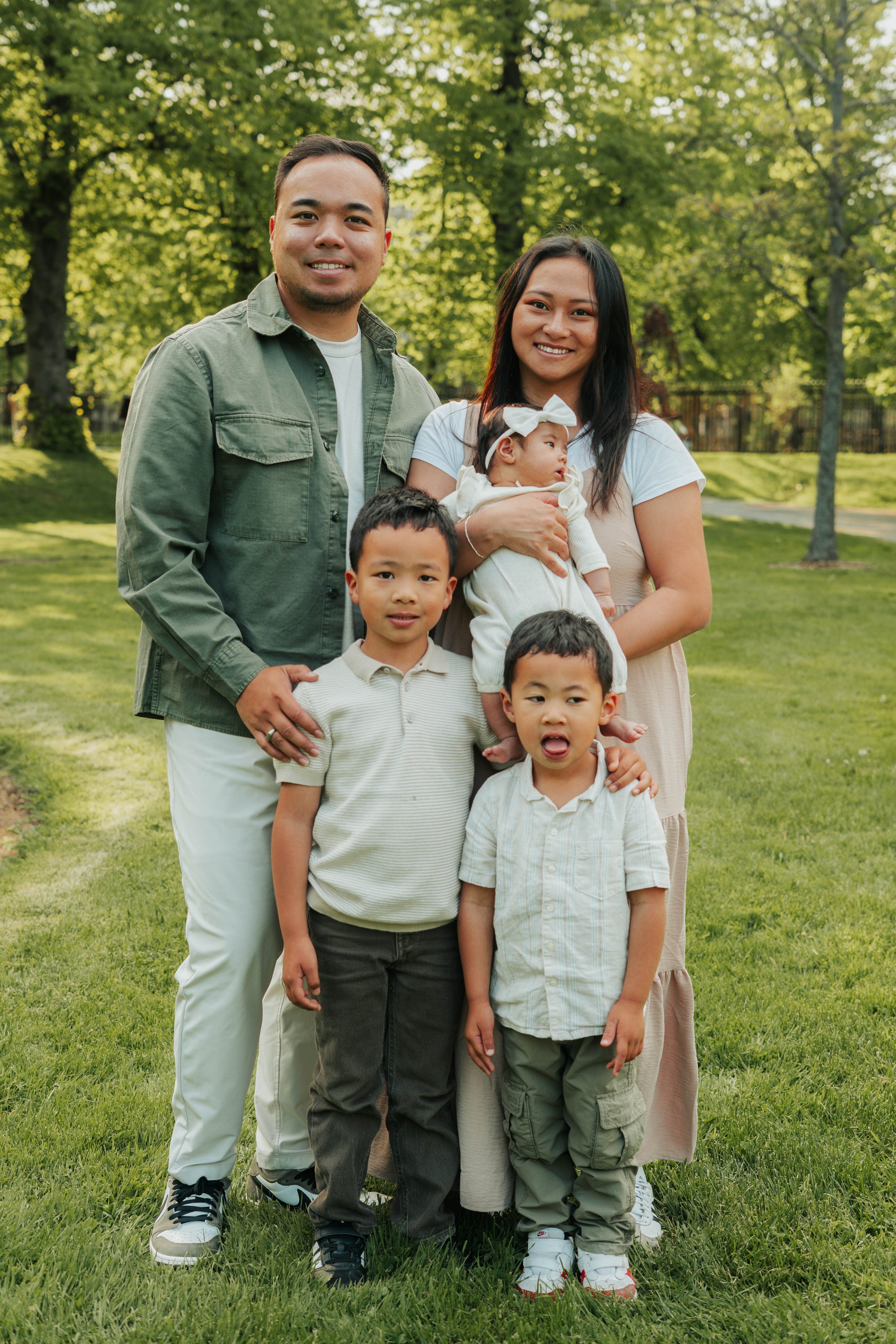 A family of six posing outdoors on a grassy area with trees in the background. The family includes two adults, two young boys, a girl, and a baby girl being held by the woman. Everyone is smiling and dressed casually.