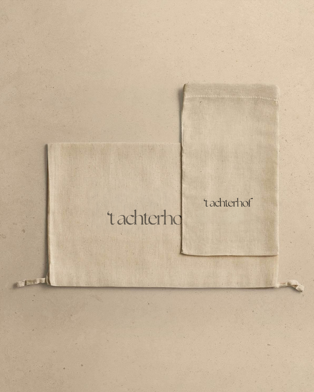 Two beige fabric pouches with the word 'achterhof' printed on them, placed on a beige surface.