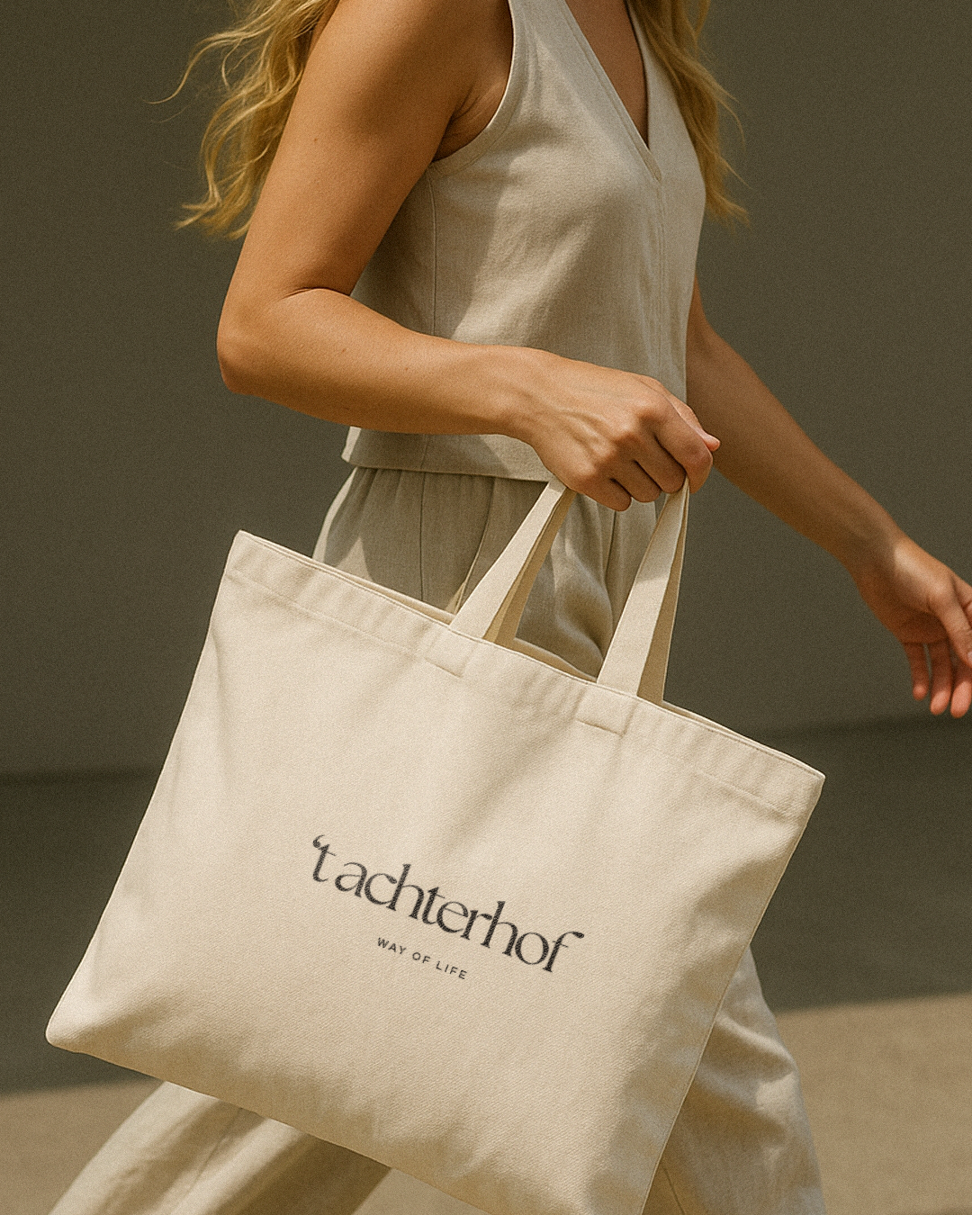 Vrouw die een beige T-shirt en broek draagt en een grote katoenen tas vasthoudt met de tekst 'tachterhof' en 'WAY OF LIFE' erop.