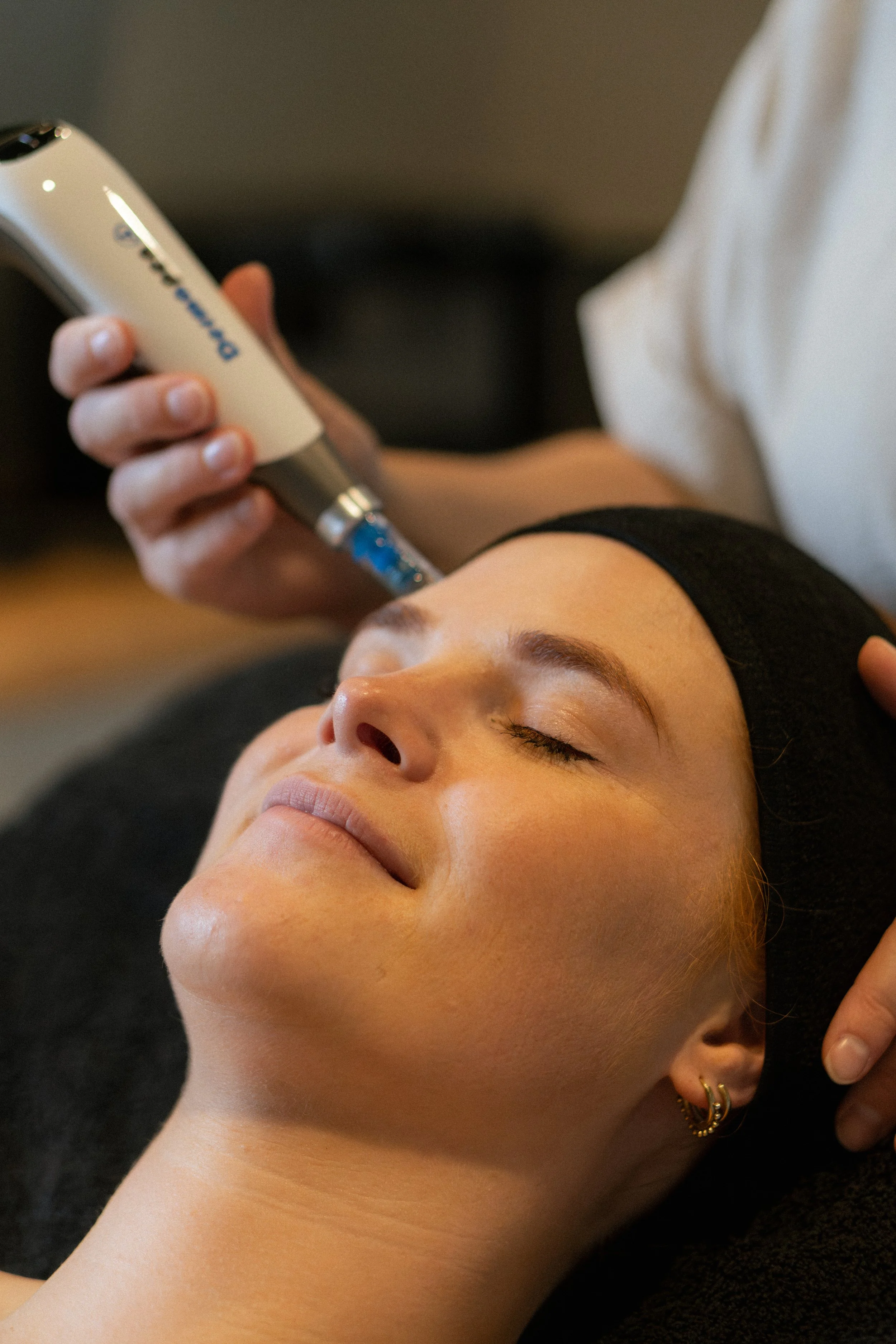 Een vrouw ligt ontspannen met haar ogen dicht terwijl een cosmetisch arts een huidbehandeling uitvoert met een apparaat op haar gezicht.