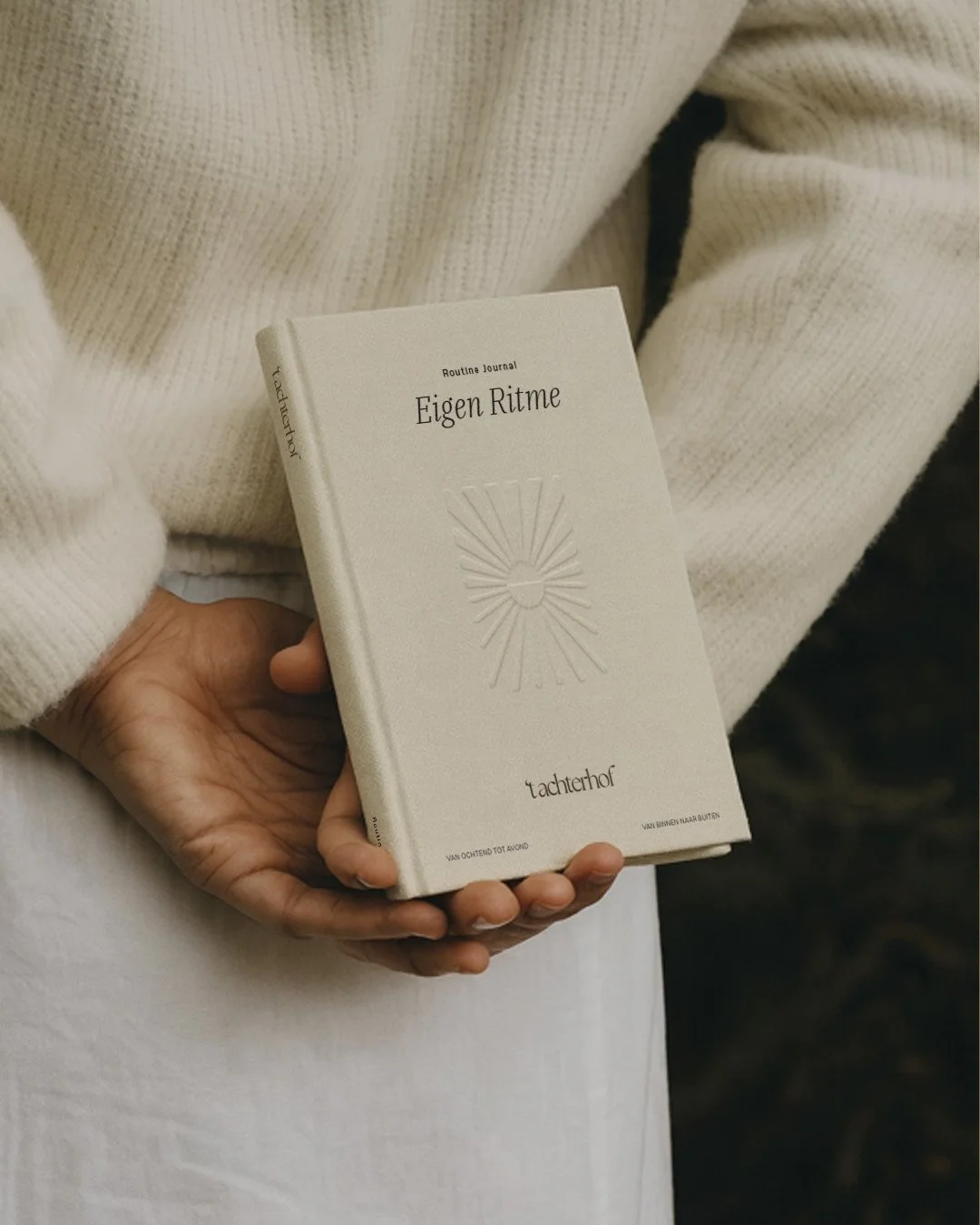 Persoon houdt een wit boek vast met de titel 'Eigen Ritme', het lijkt een dagboek of notitieboek te zijn, met een hand die het vasthoudt, omgeven door een witte trui en lichte kleding.