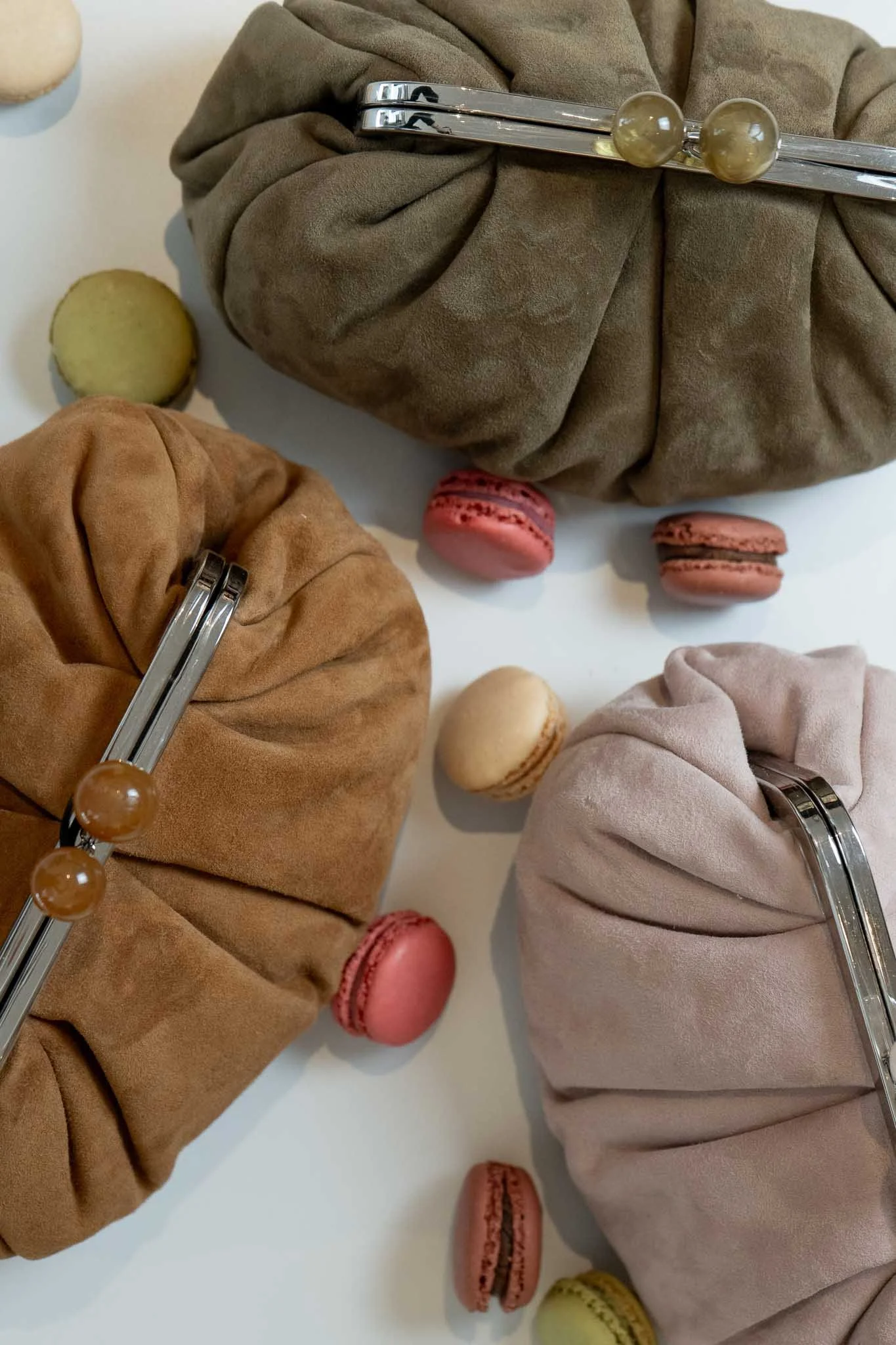 Three fabric clutch purses in beige, brown, and taupe colors with metal and pearl clasps on a white surface, surrounded by colorful macarons in pink, green, yellow, and beige.
