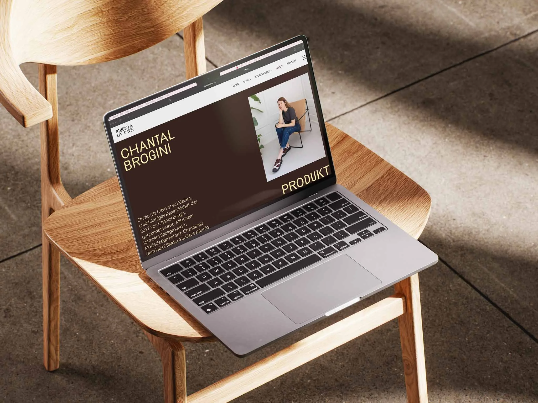 A silver MacBook laptop displaying a website, placed on a small wooden chair with a beige fabric seat, outdoors on a concrete surface.