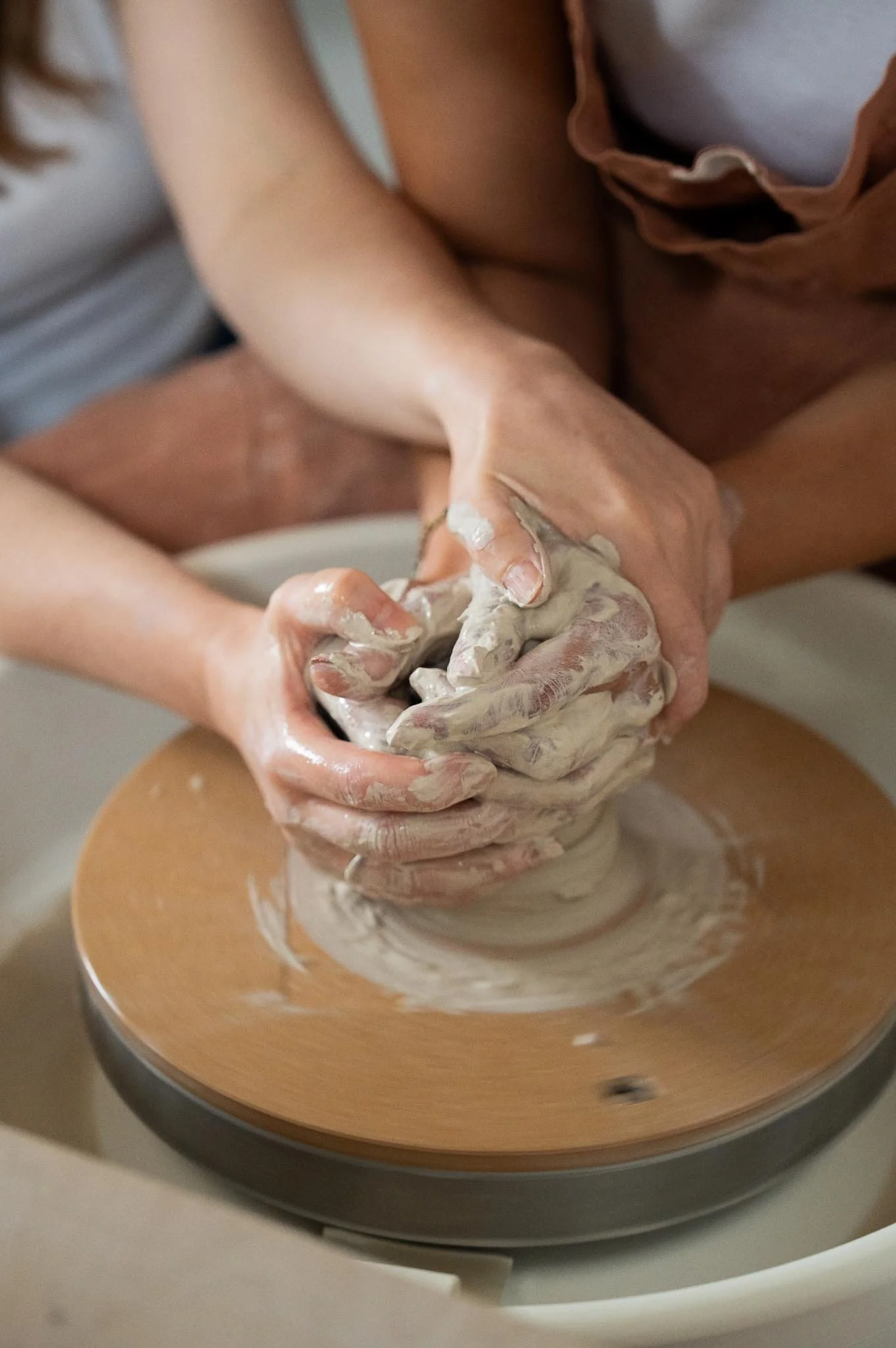Two people are shaping clay on a pottery wheel, their hands covered in wet clay.
