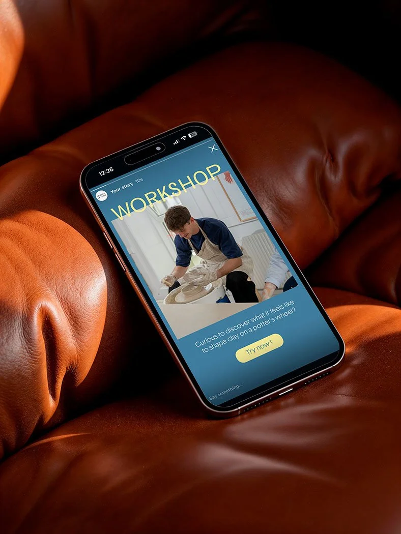A smartphone lying on a brown leather couch displays a social media story about a workshop that involves shaping clay on a potter's wheel, with a picture of a young man working with clay.