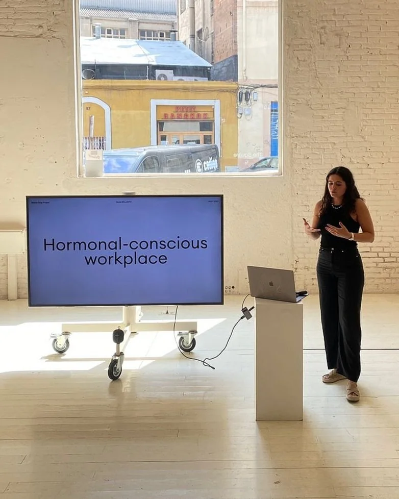 A woman is giving a presentation about a 'hormonal-conscious workplace' in a bright room with large windows. There is a large screen on wheels displaying the presentation, and a laptop on a small stand next to her.