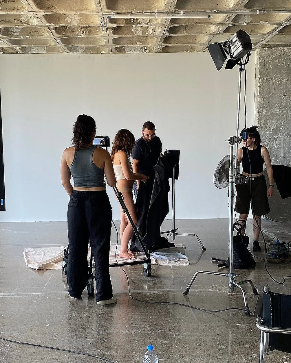 A photoshoot setup with a woman standing on a platform, surrounded by crew members and equipment in a studio with a white background and unfinished ceiling.