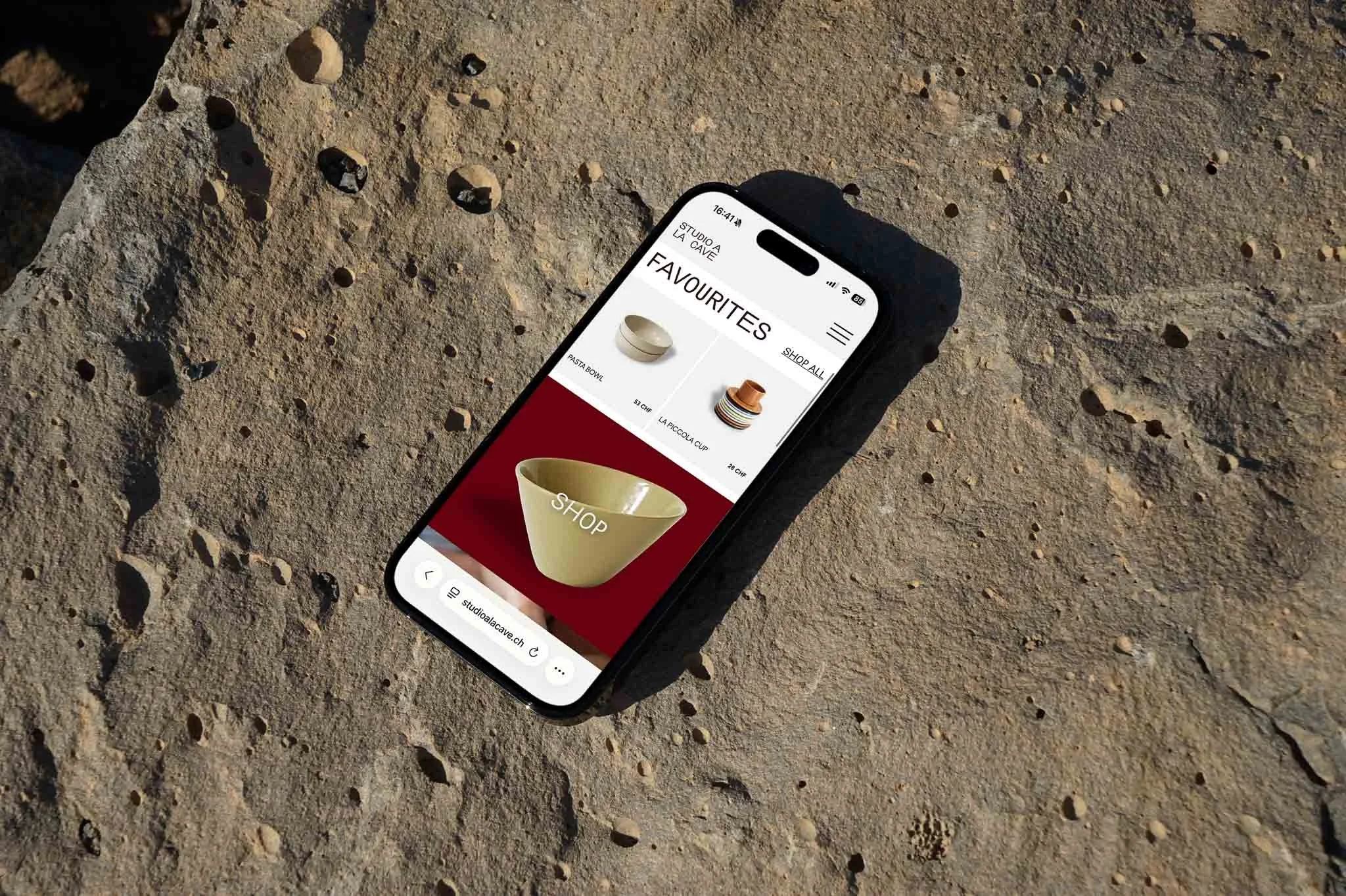 A smartphone lying on a rough, sandy surface displays an online shop for ceramic bowls and cups, with the word "SHOP" featured prominently on a beige bowl.
